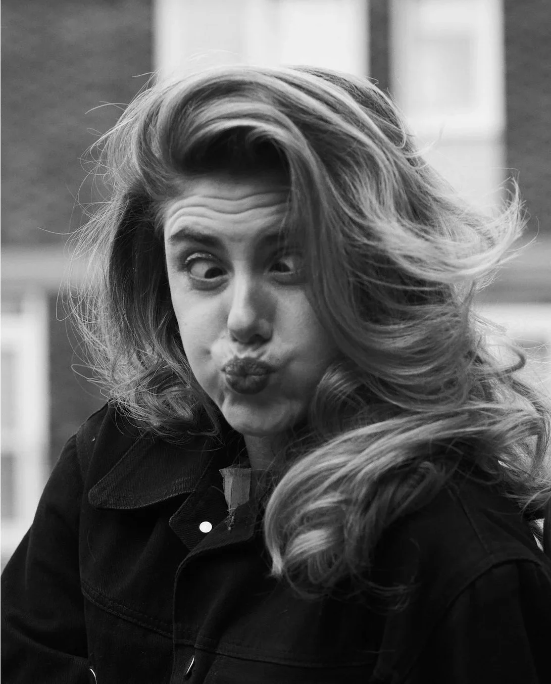 A woman with long, wavy hair making a playful face with pursed lips and wide eyes, in black and white.