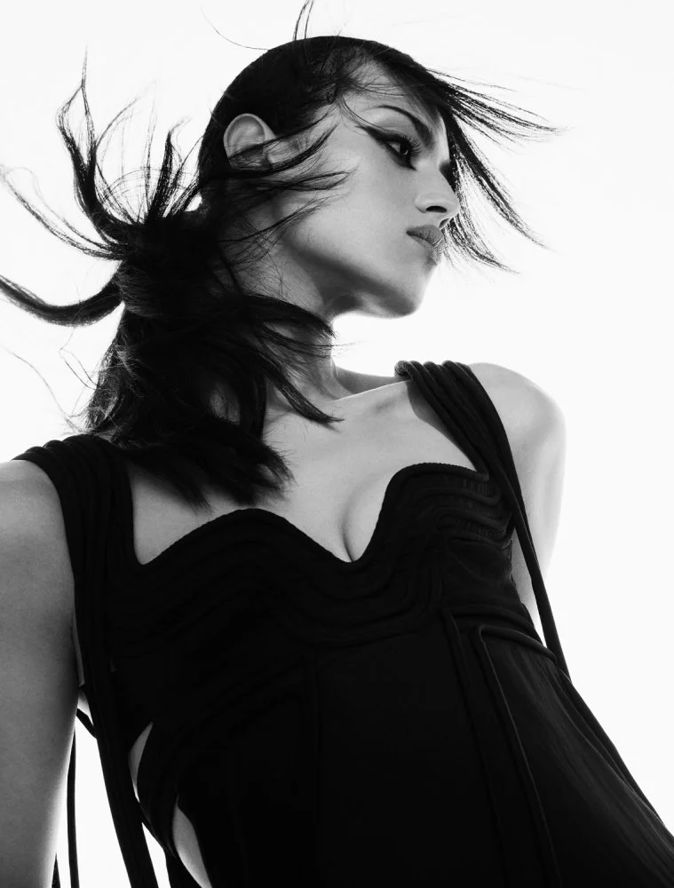 Black and white side profile of a woman with windblown hair wearing a black sleeveless dress, against a plain background.