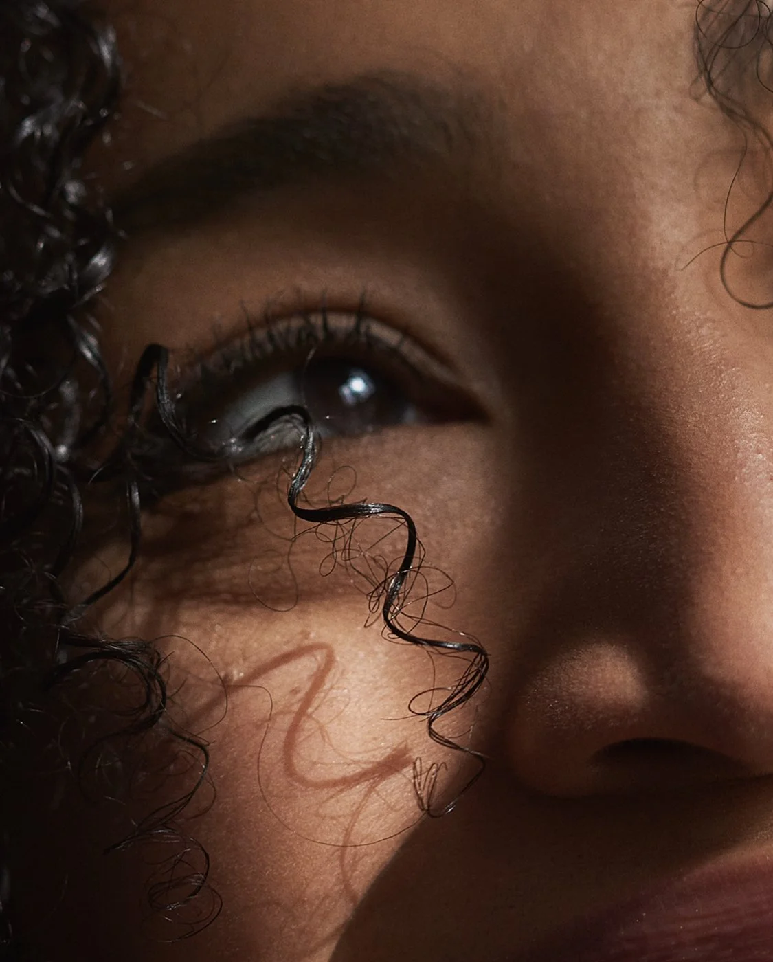 A close-up of a person's face, showing the eye, curly hair, and skin texture.