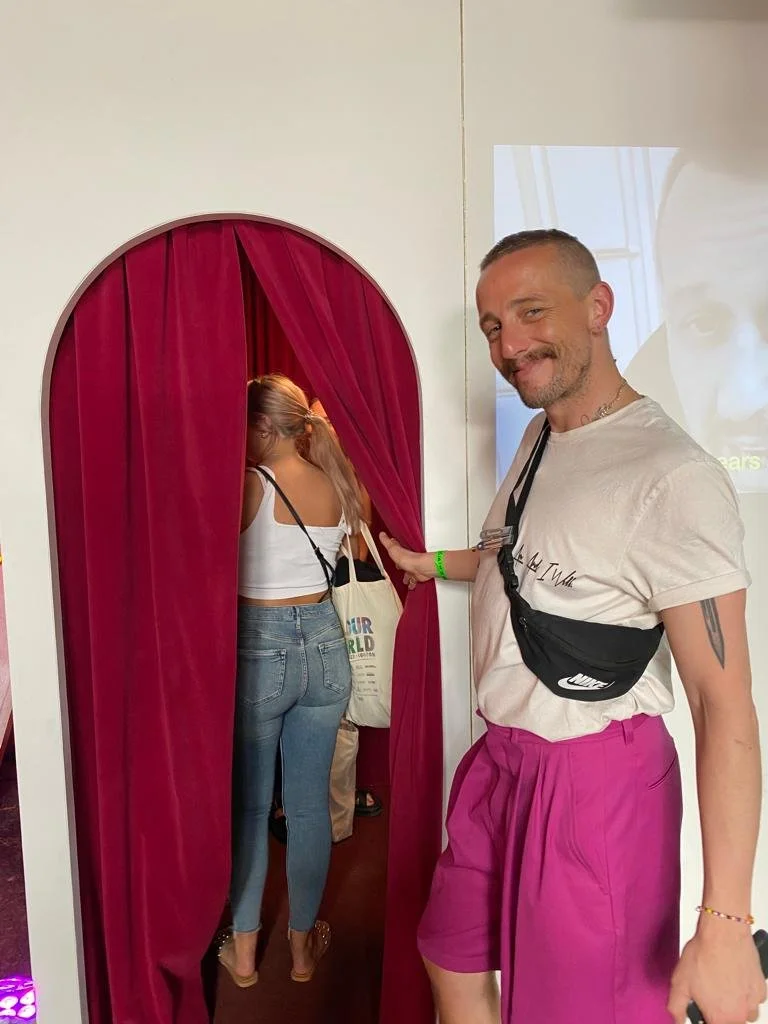 A man smiling and wearing pink shorts, a white T-shirt, and a black Nike fanny pack is pulling back a red curtain, revealing a woman with long hair in a ponytail, wearing a white crop top and blue jeans, entering a private area. The scene takes place