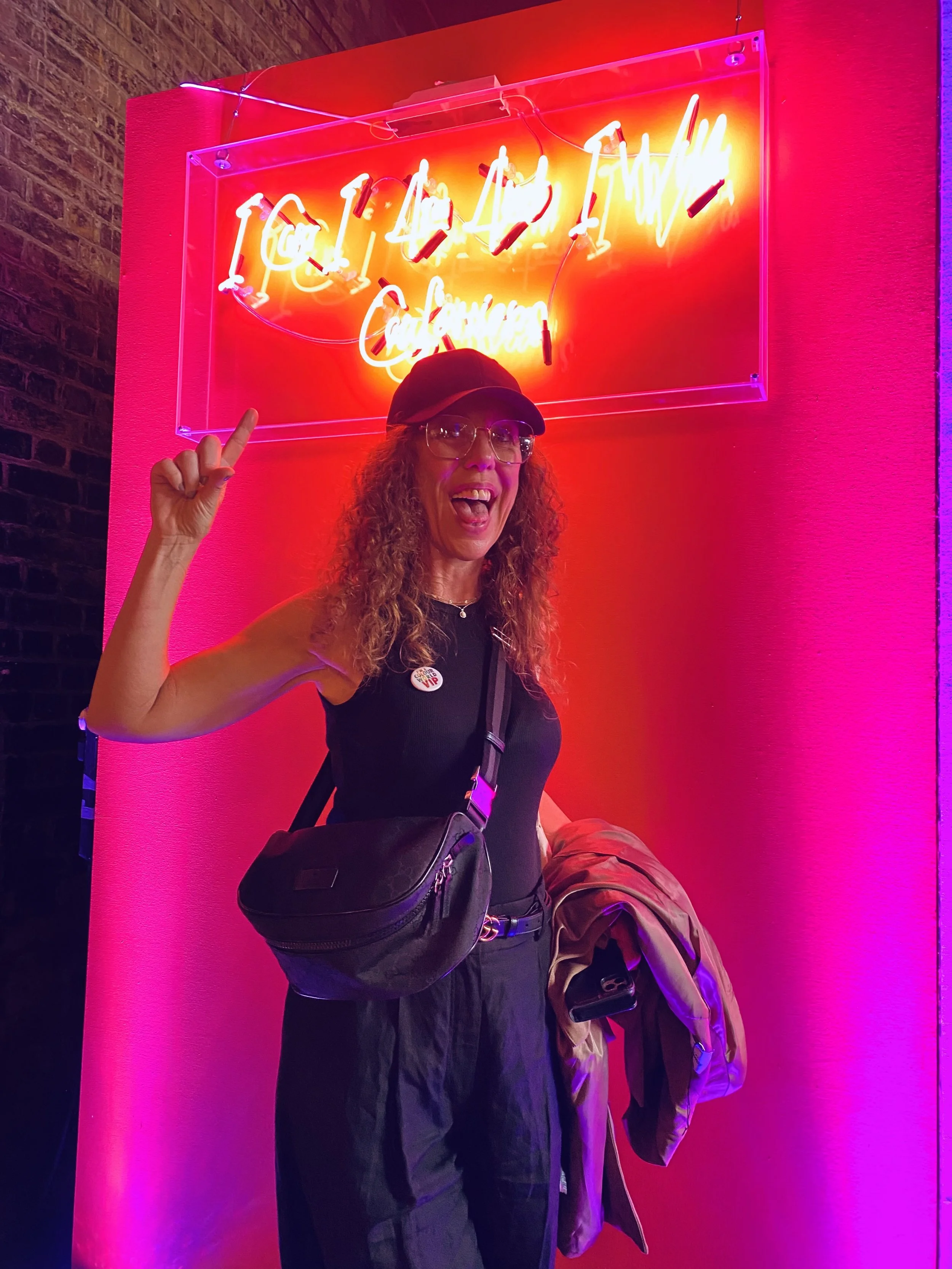 A woman with curly hair, glasses, wearing a black cap and tank top, standing in front of a bright pink wall with a colorful neon sign, holding a jacket and a bag, smiling and pointing up.