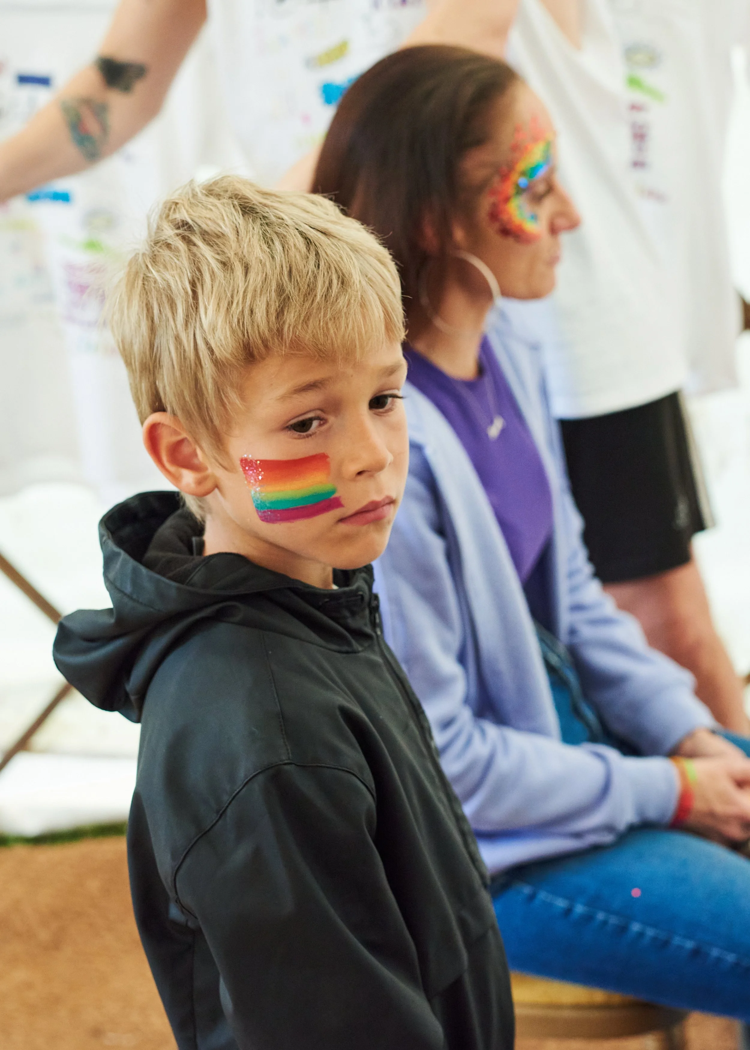 A young boy with blonde hair and rainbow face paint is sitting with a serious expression. Behind him, an adult woman with rainbow face paint is seated, wearing a purple shirt and a light blue jacket. Several other people with rainbow face paint and w