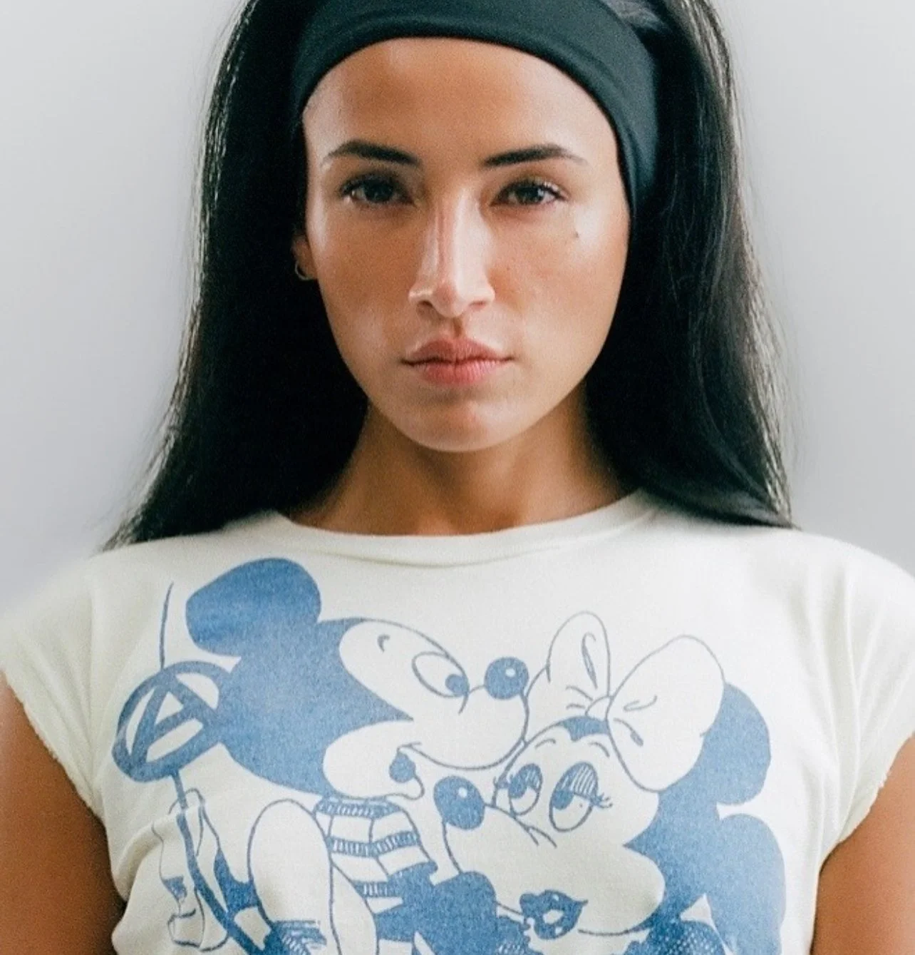 A woman with long dark hair, wearing a black headband and a white T-shirt featuring a blue graphic of Mickey Mouse and Minnie Mouse.