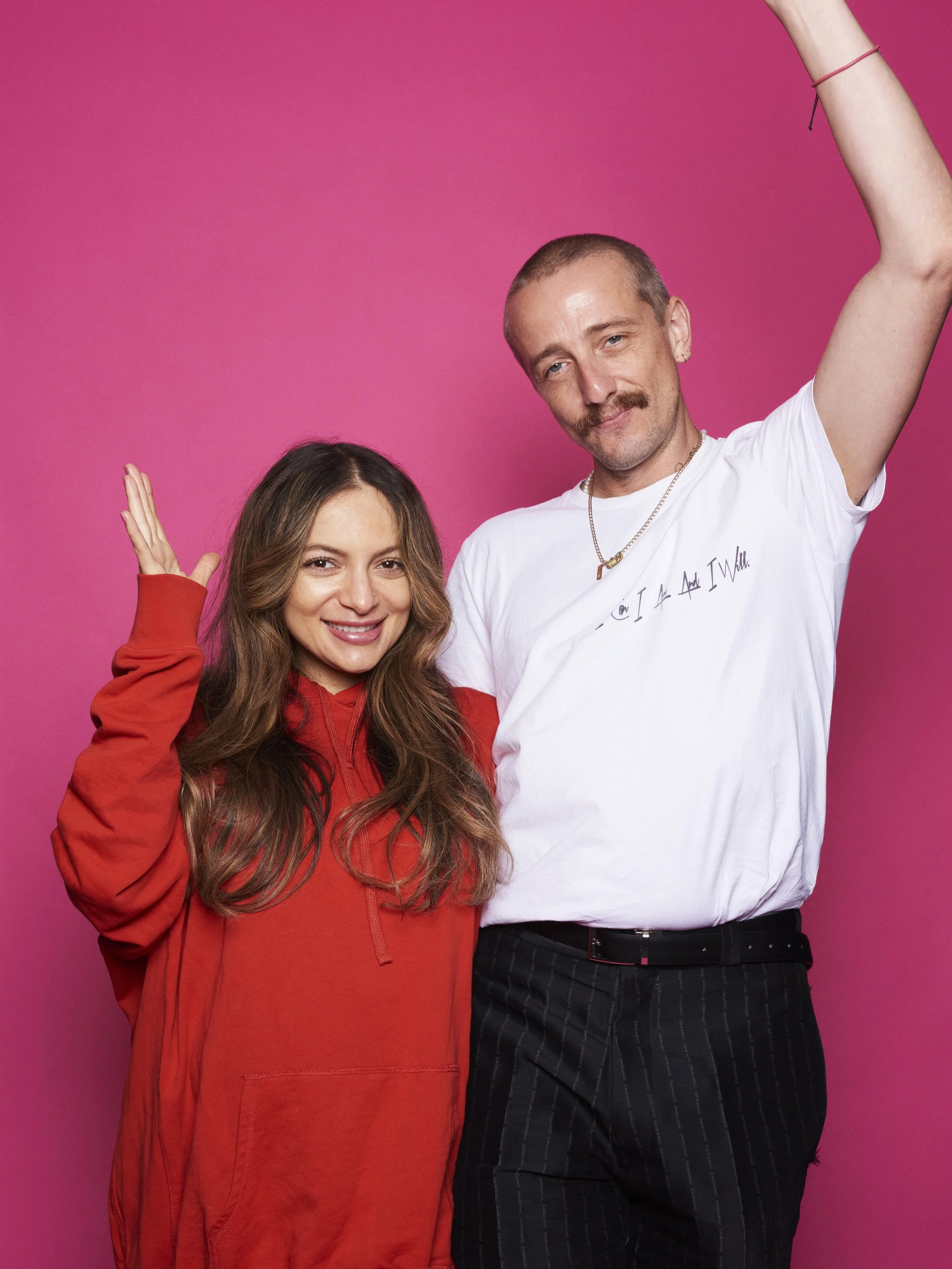 A smiling woman in a red hoodie standing next to a man with a mustache, wearing a white t-shirt and striped pants, against a pink background.