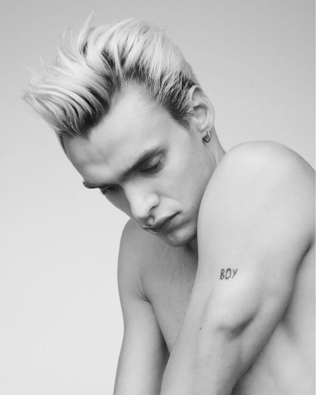 Black and white photo of a young man with platinum blonde hair looking down, shirtless with a tattoo that says 'BOY' on his arm, wearing earrings.