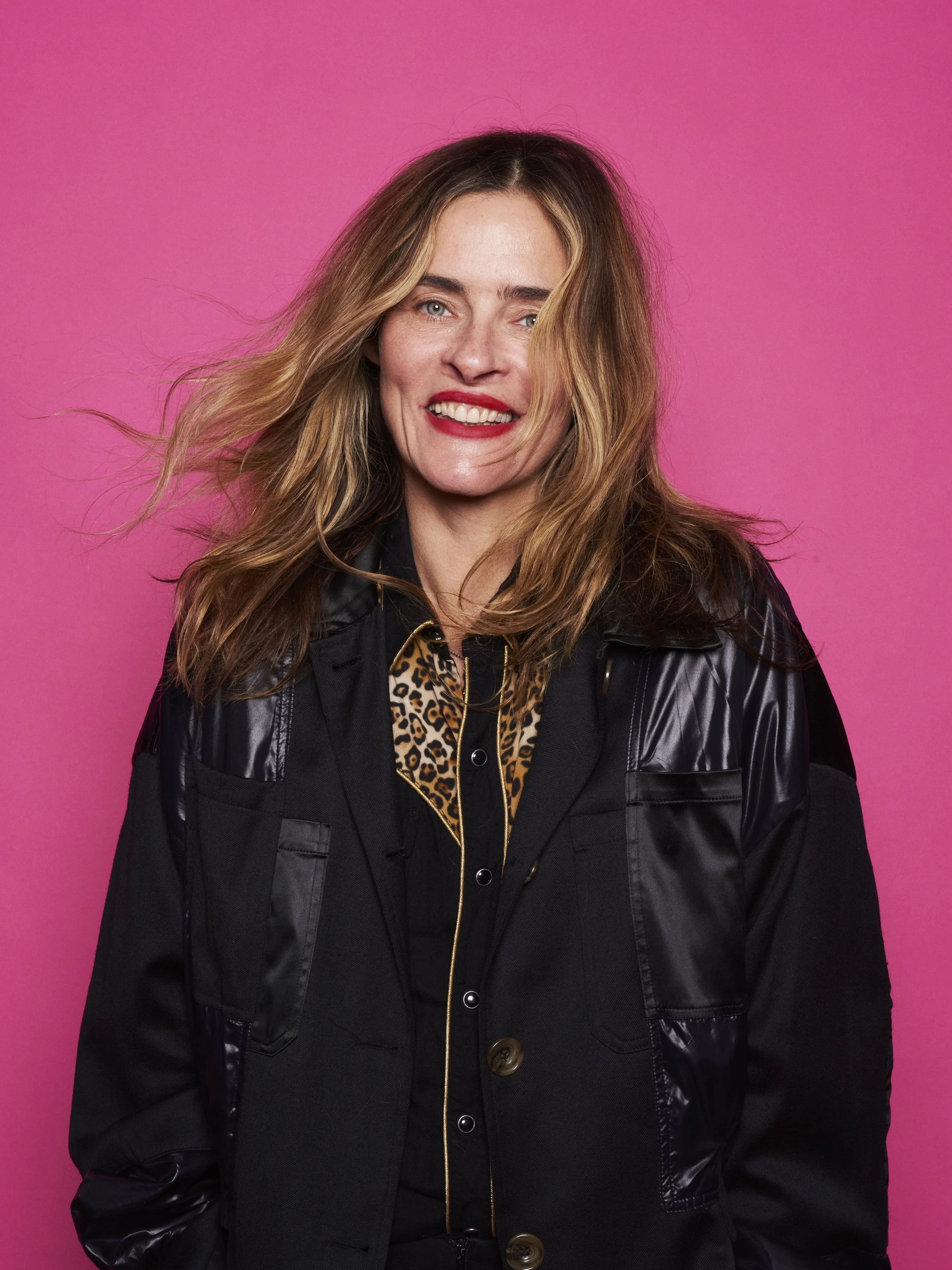 A woman with long, wavy blonde hair smiling against a pink background, wearing a black jacket and a leopard print shirt.