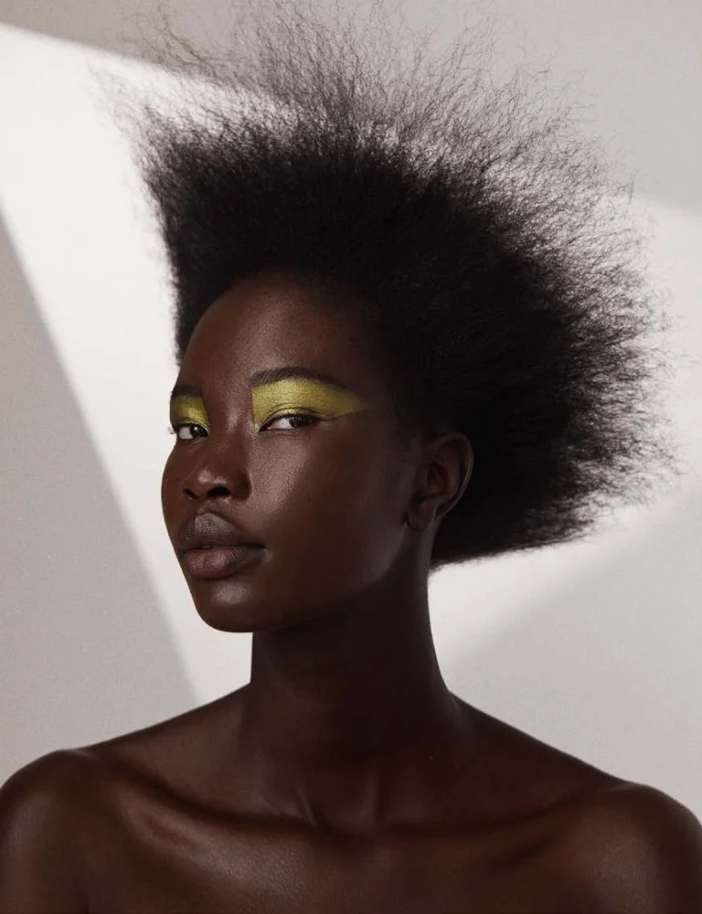 A young woman with dark skin and natural textured hair, wearing bright yellow eyeshadow, facing slightly left, with a neutral expression against a plain background.