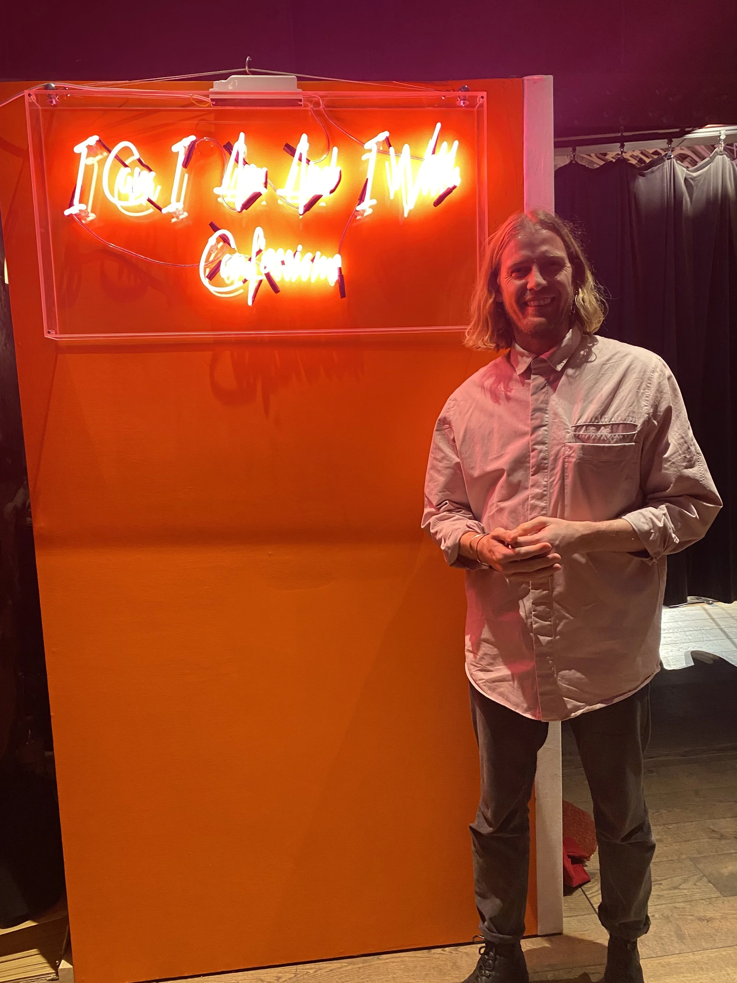 A man with long blond hair and a beard, wearing a light-colored button-up shirt and dark pants, stands smiling in front of an orange wall with a neon sign that reads 'I Can Tame My Crying' in a bright, glowing script.