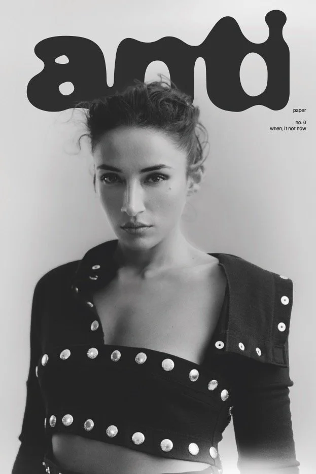 Black and white photo of a woman with her hair tied up, wearing a jacket with metallic studs, standing against a plain background. The word 'and' is written in large, stylized letters at the top of the image, with small text reading 'paper no. 0 when