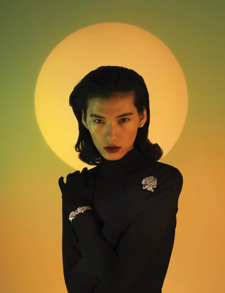 A woman with dark hair in a black turtleneck sweater standing in front of a yellow circle/backdrop, with serious expression, dark lipstick, and wearing decorative jewelry.