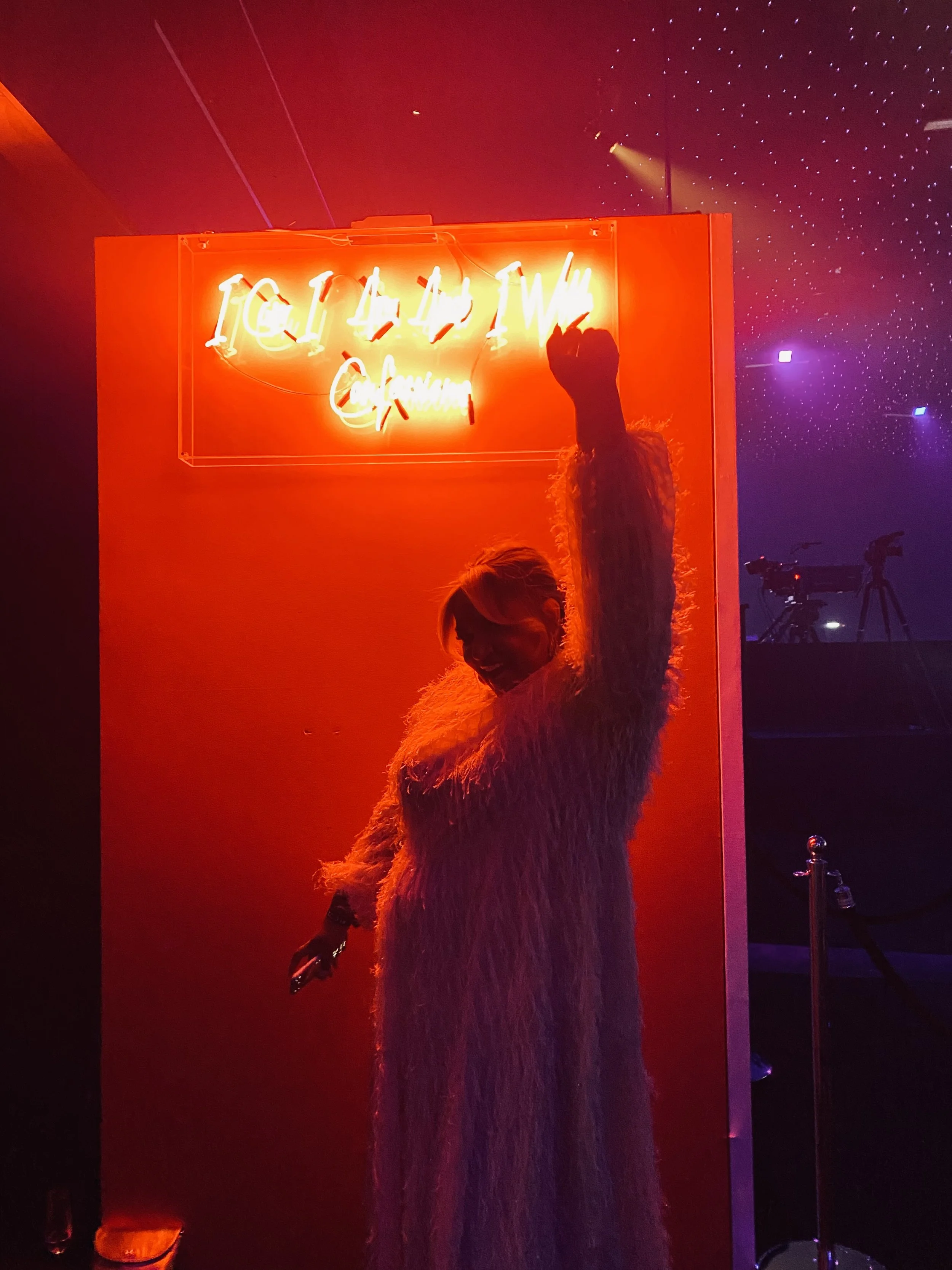 Woman in a long, furry coat standing in front of a red neon sign that says 'TICKLE ME GORGEOUS', with her arm raised and holding a small object in her hand, inside a dimly lit venue with a starry ceiling and cameras in the background.