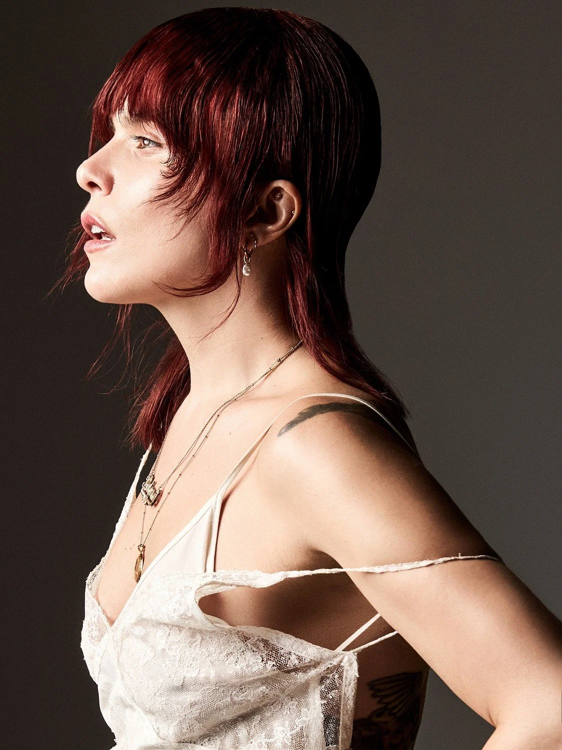 Profile of a woman with short, red hair, wearing delicate jewelry, and a lace, spaghetti strap top against a dark background.