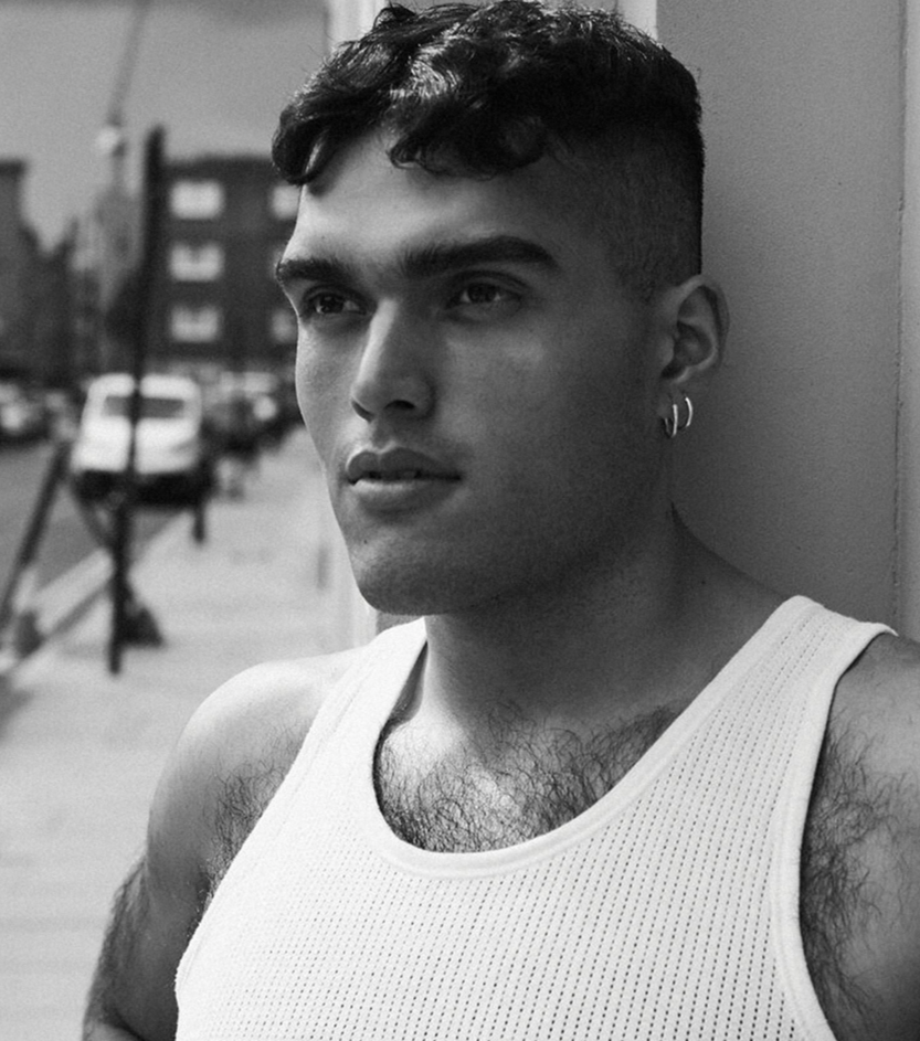 A young man with short dark hair and hoop earrings wears a sleeveless athletic shirt, leaning against a wall on an urban street, with parked cars and buildings in the background, in black and white.