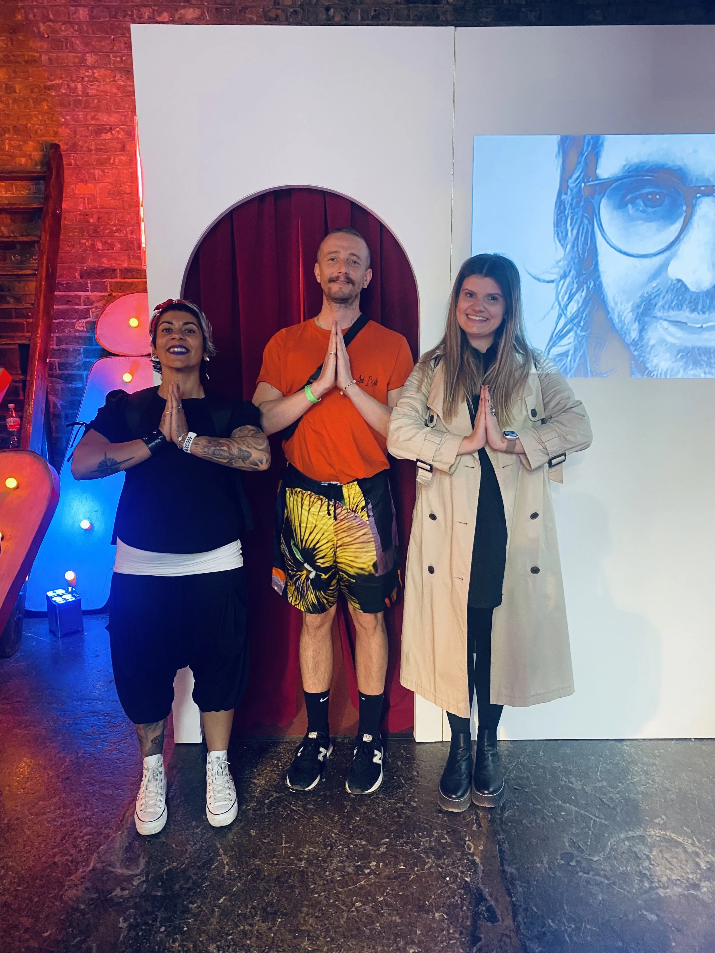 Three people standing together in a room, posed with hands in prayer position, smiling. They are in front of a red curtain and a partial portrait on the wall. Colorful lights illuminate the background.