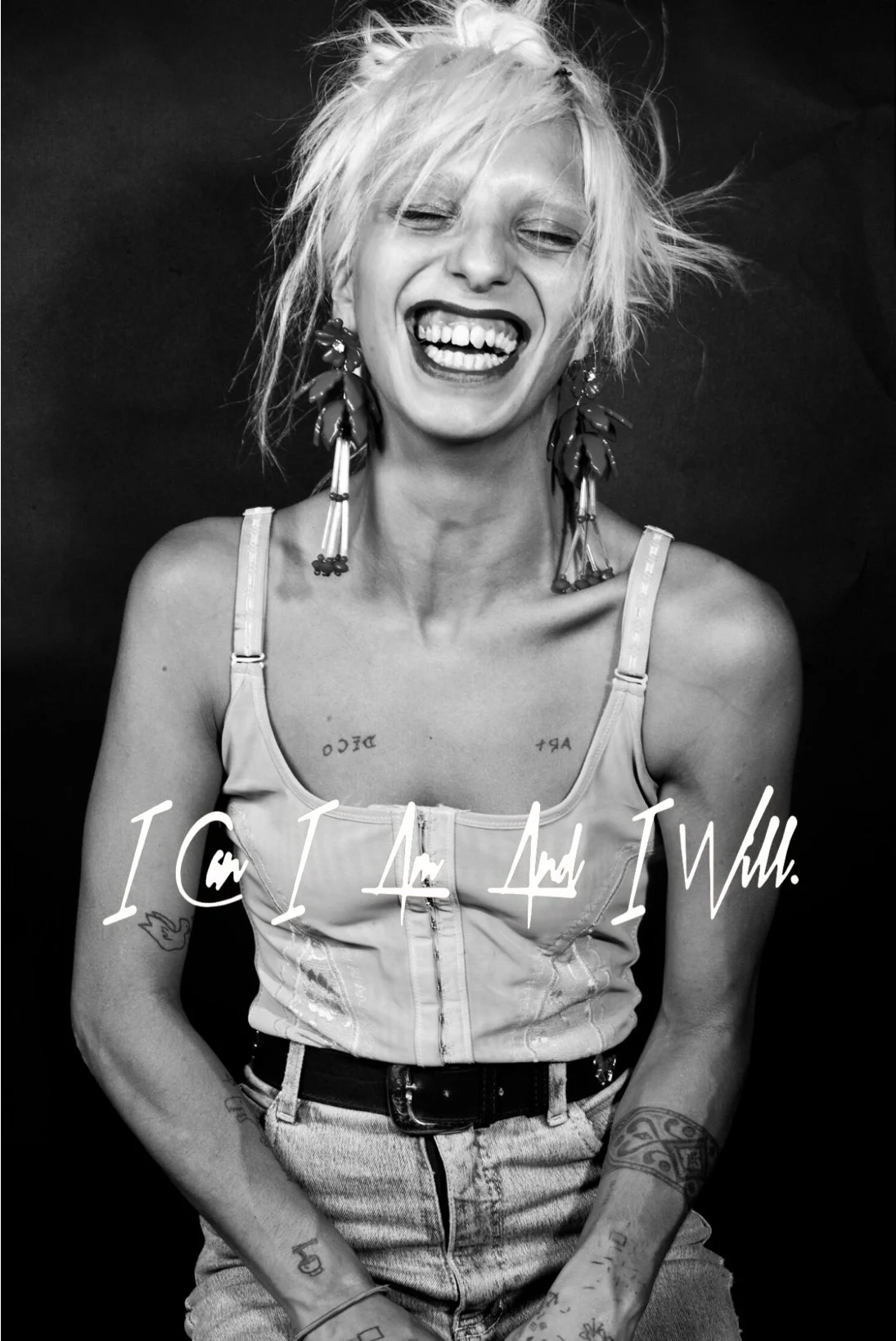 A woman with short, messy platinum blonde hair smiling and laughing. She is wearing large, leaf-shaped earrings, a light-colored tank top, and high-waisted jeans. She has visible tattoos on her arms and chest, and there is white cursive text overlayi