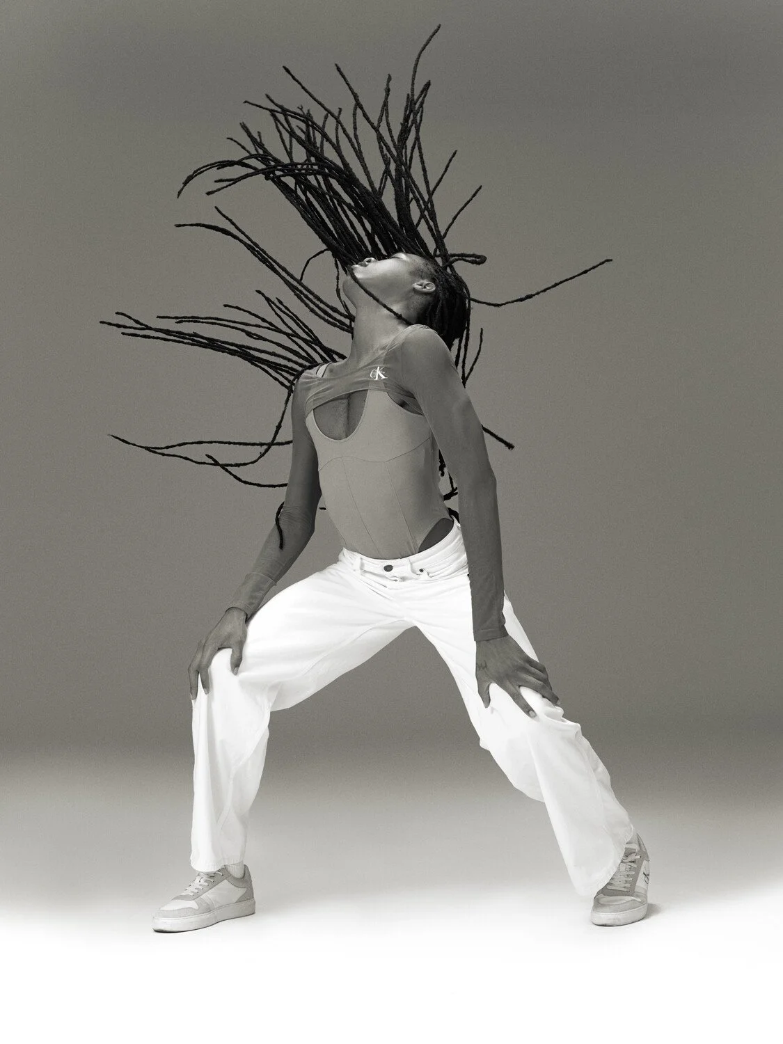 A woman with long dreadlocks extending outward, wearing a sleeveless top and white pants, poses in a wide stance against a plain background.