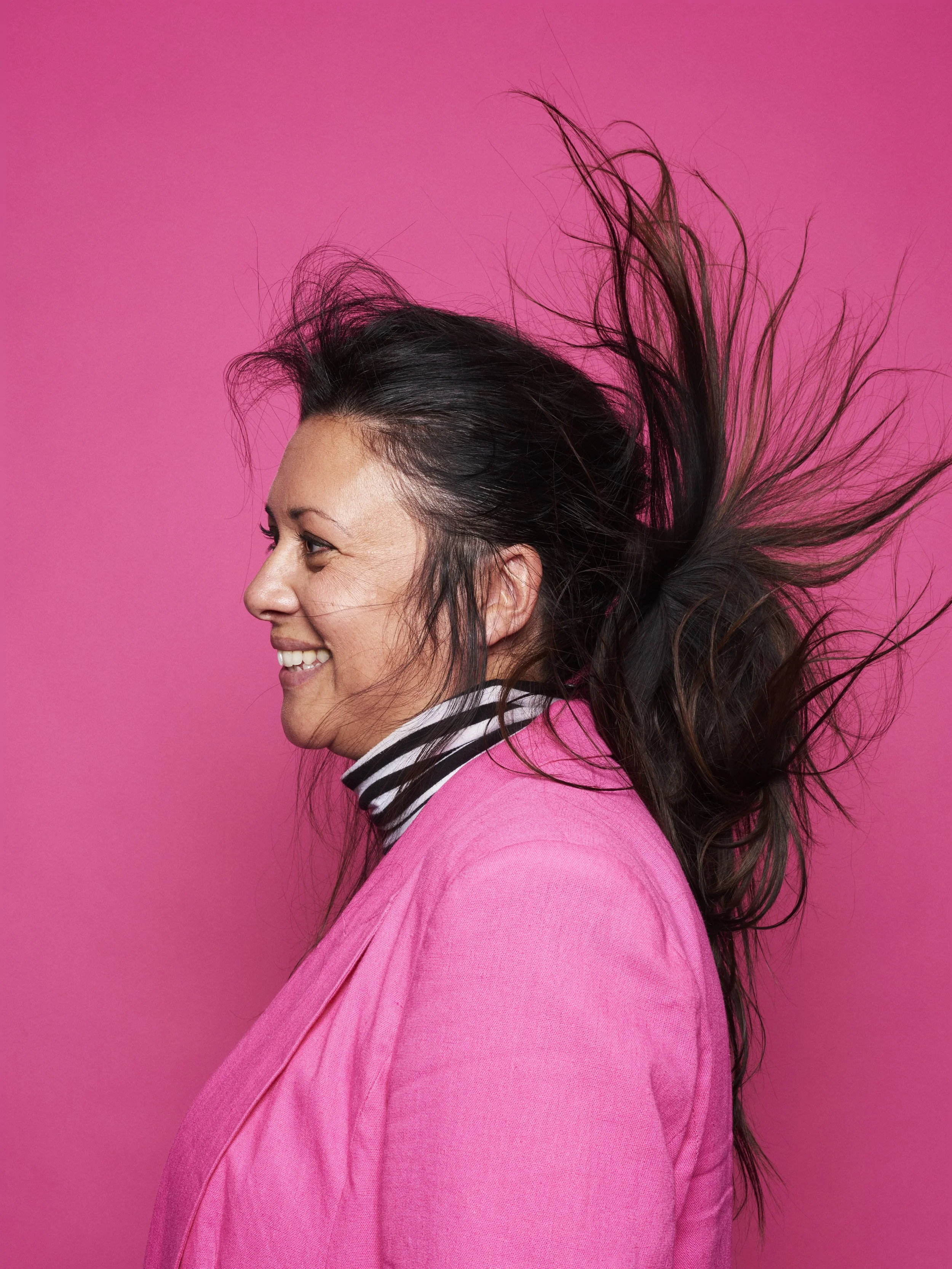 Side profile of a smiling woman with long dark hair, wearing a pink blazer over a striped turtleneck, against a pink background.