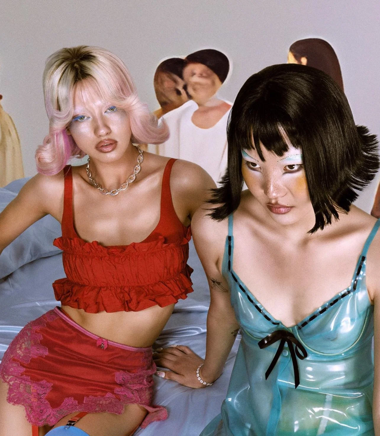 Two women sitting on a bed, one with pink hair wearing a red ruffle top and pink lace skirt, the other with black hair in a blue satin dress with black ribbons. In the background, there are blurred humorous images of people and a woman with short hai