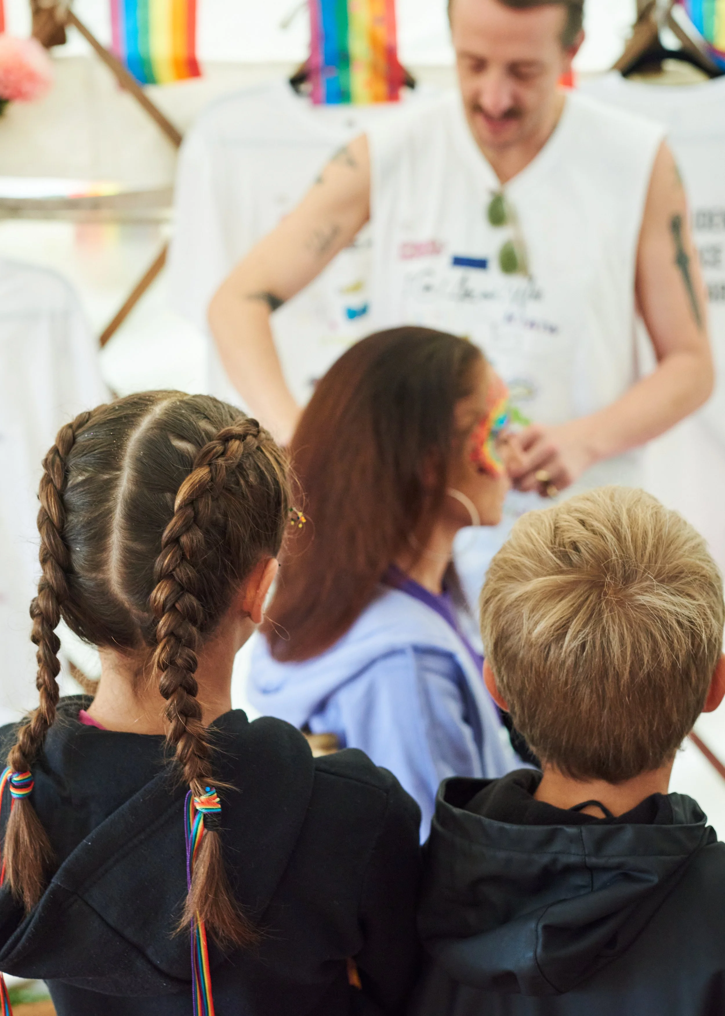 A rainbow-themed face painter painting a rainbow on a woman's face at an outdoor event, with children watching.