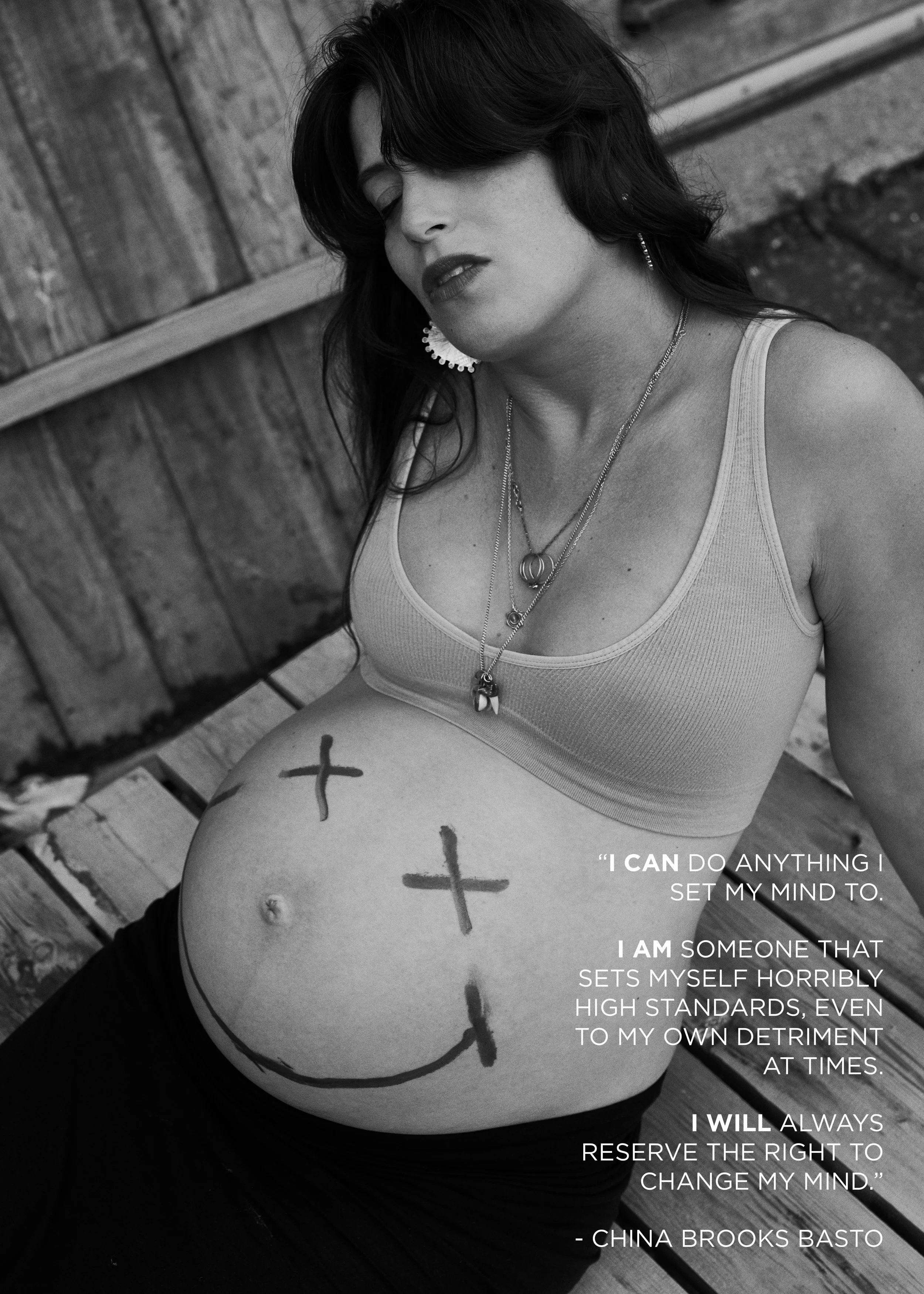 A black and white photo of a pregnant woman with dark hair, wearing a sports bra and sitting outdoors on a wooden bench. She has hand-drawn crosses and a smiley face on her belly, and is wearing multiple necklaces and earrings. The image contains a q