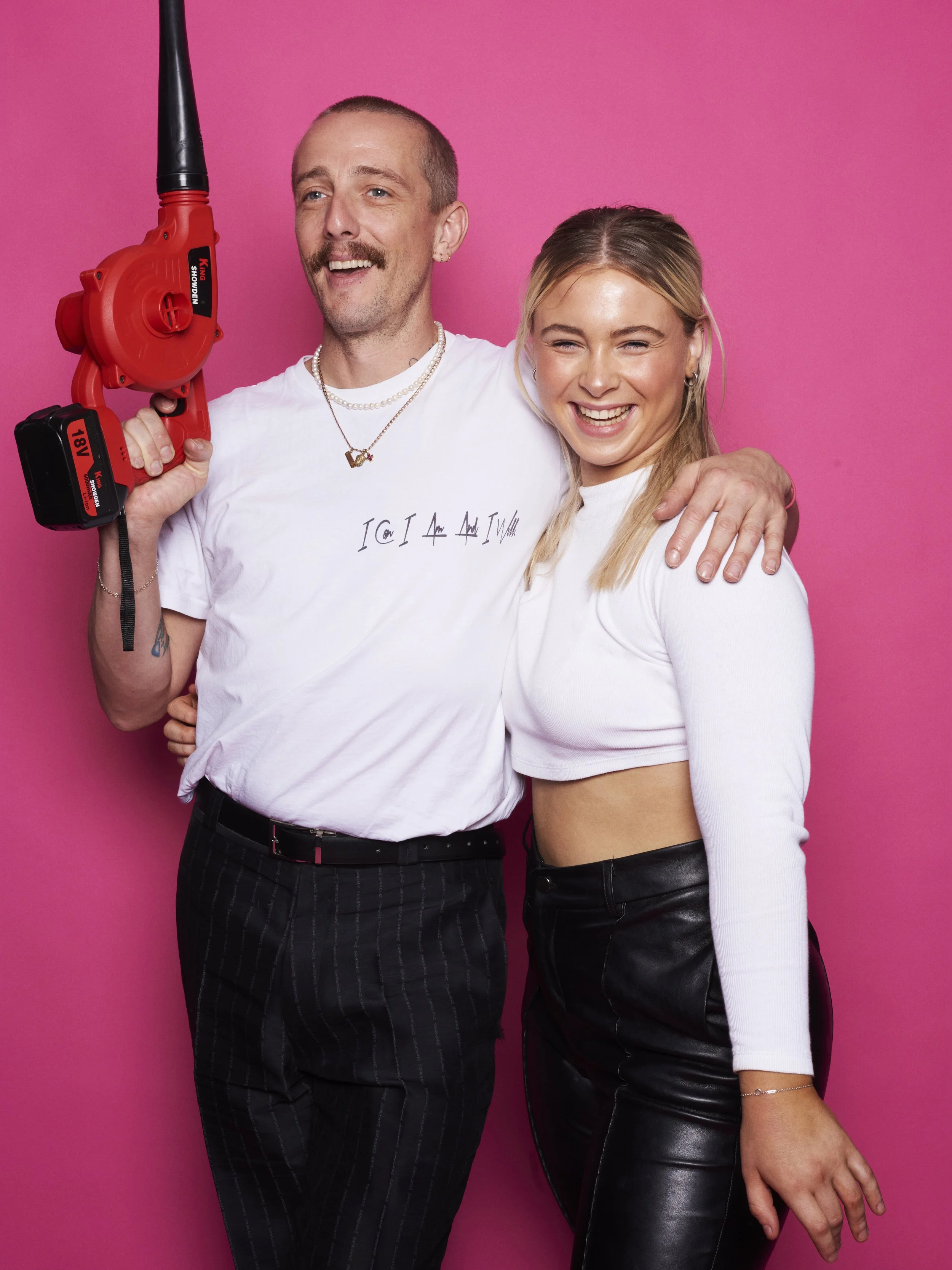 Two happy people, a man and a woman, standing together against a pink background. The man is holding a red power tool and has his arm around the woman. They are both smiling.