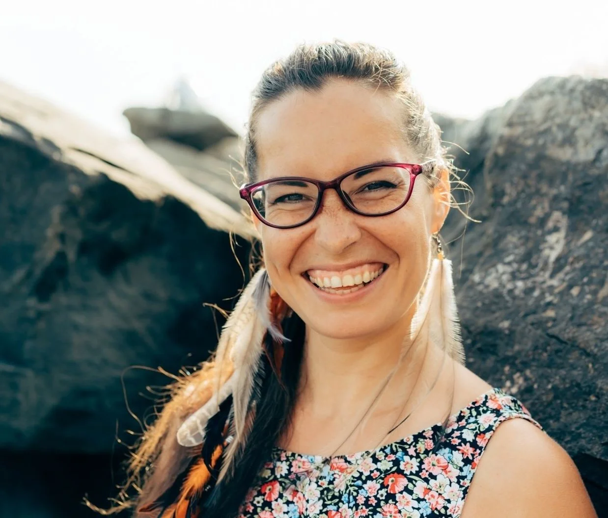 Een lachende vrouw met bruin haar, gekleed in een bloemenjurk, draagt kleine glazen en oorbellen, met rotsen op de achtergrond.