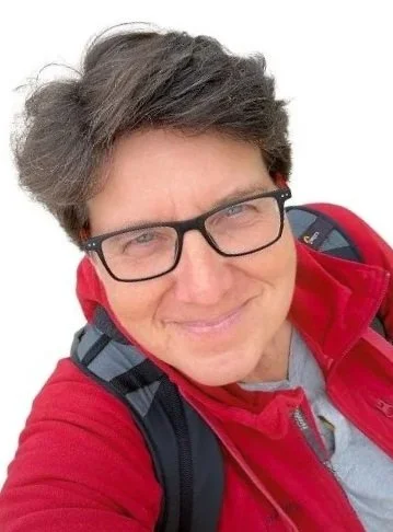 Persoon met kort bruin haar, zwarte bril, lachend, rode jas en rugzak op een witte achtergrond.