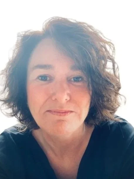 Een vrouw met schouderlange, golvende bruin haar en blauwe ogen, gekleed in een zwart shirt, lachend op een lichte achtergrond.