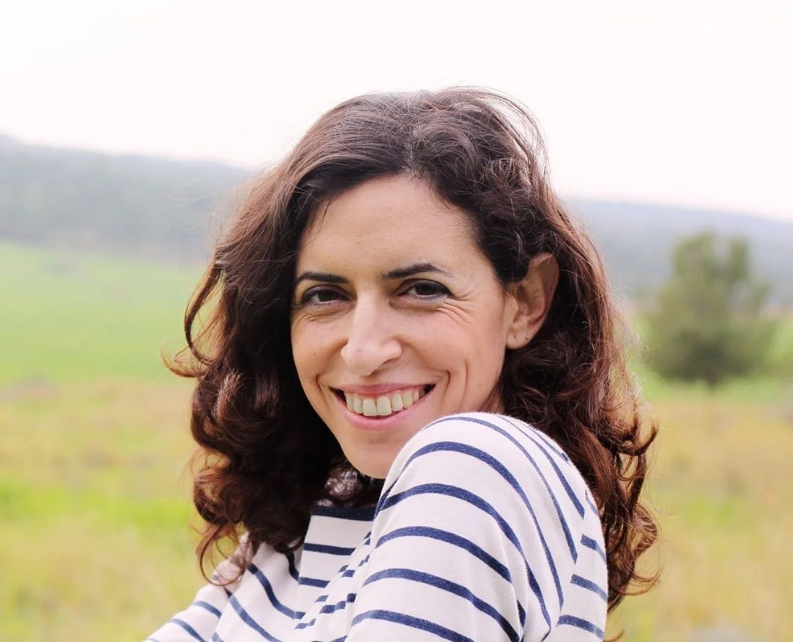 Gelukkige vrouw met bruin, krullend haar, zittend buiten op een groene velden, glimlachend naar de camera.