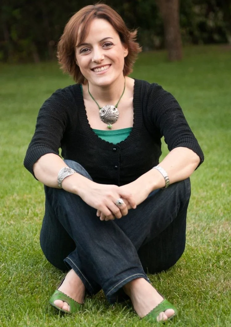 vrouw met kort bruin haar zittend op gras, lachend, in een park, gekleed in een zwarte cardigan, groene top, blauwe jeans, groene schoenen, met sieraden, en een grote ronde hanger om haar hals.