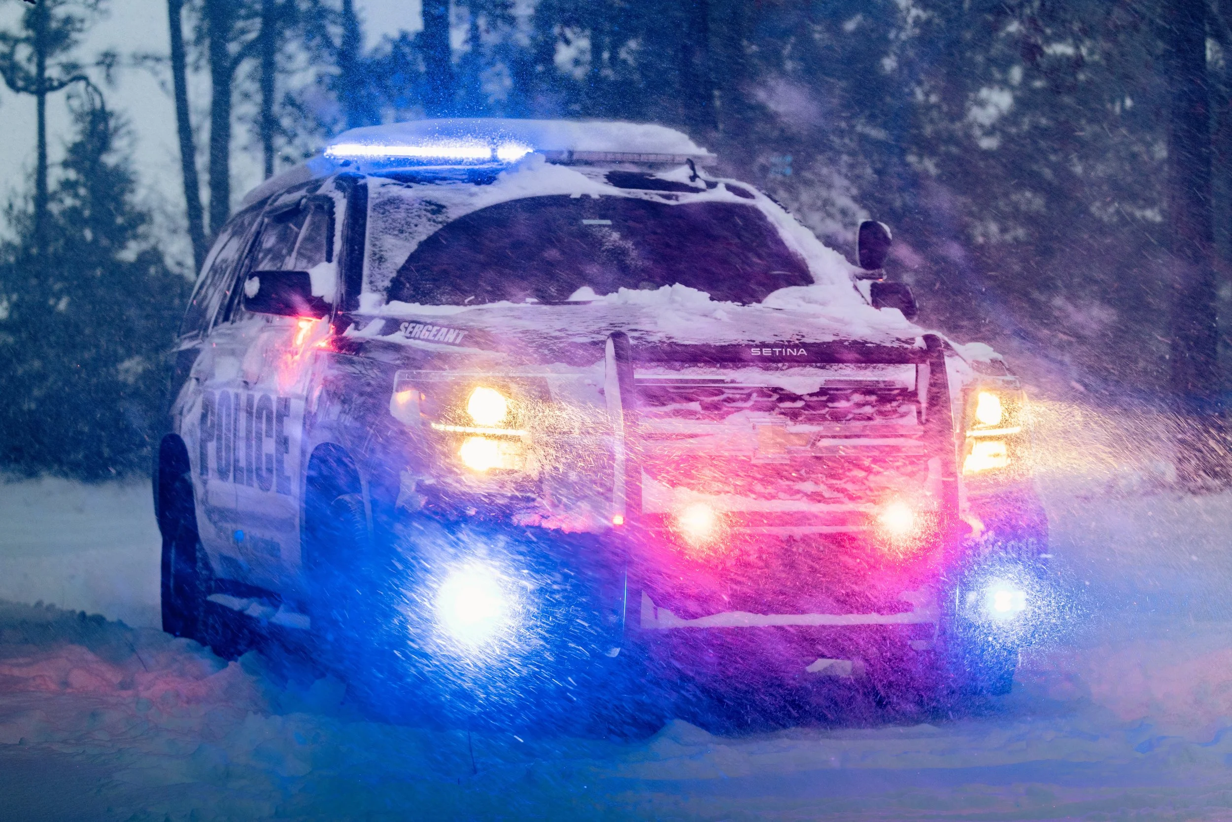 A police SUV with flashing lights driving through a snowy landscape at night, surrounded by snow-covered trees.