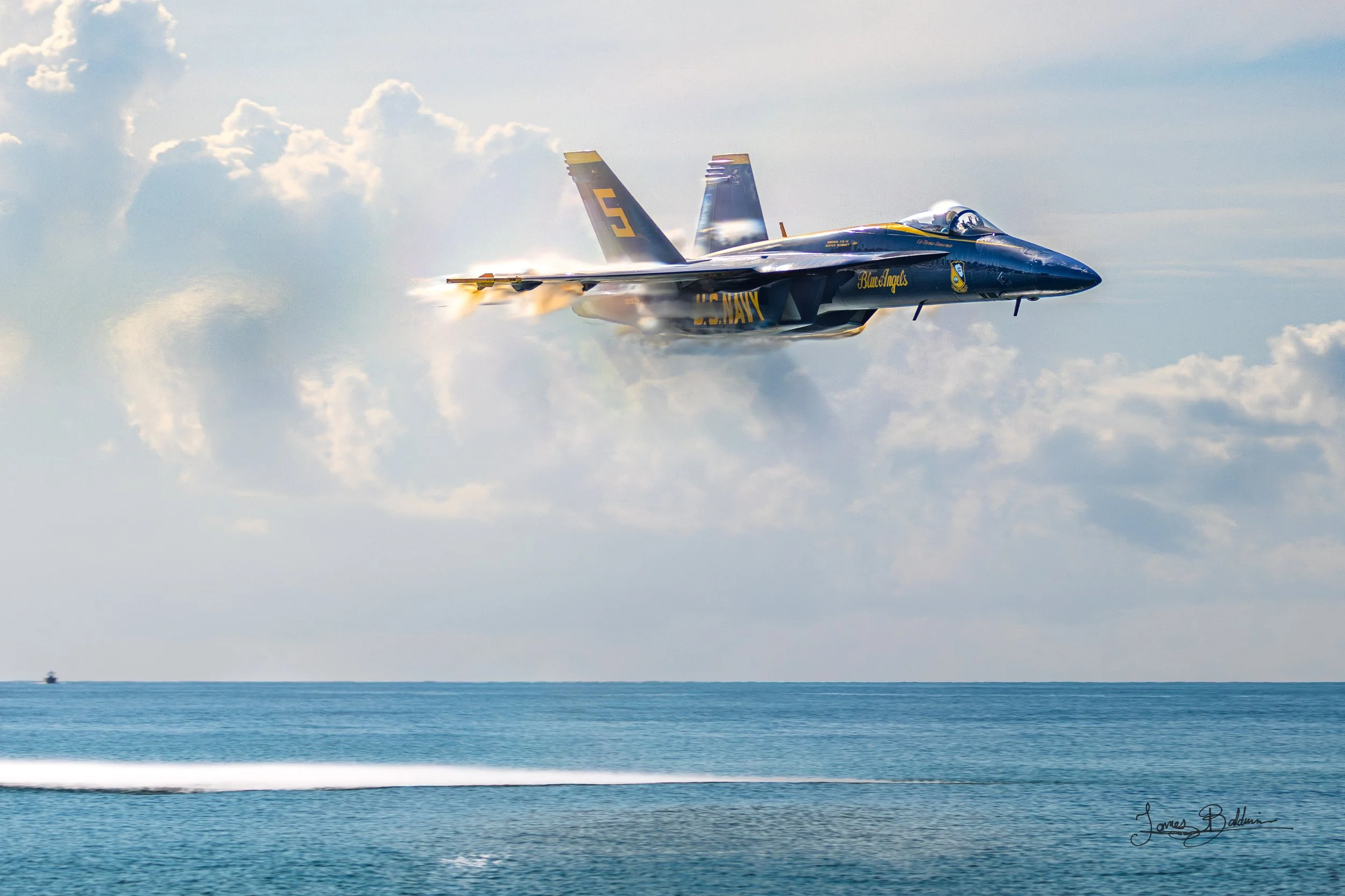 A fighter jet flying low over the ocean, leaving a vapor trail behind. The jet is painted in navy colors with yellow accents and markings, and has a shark teeth nose design.