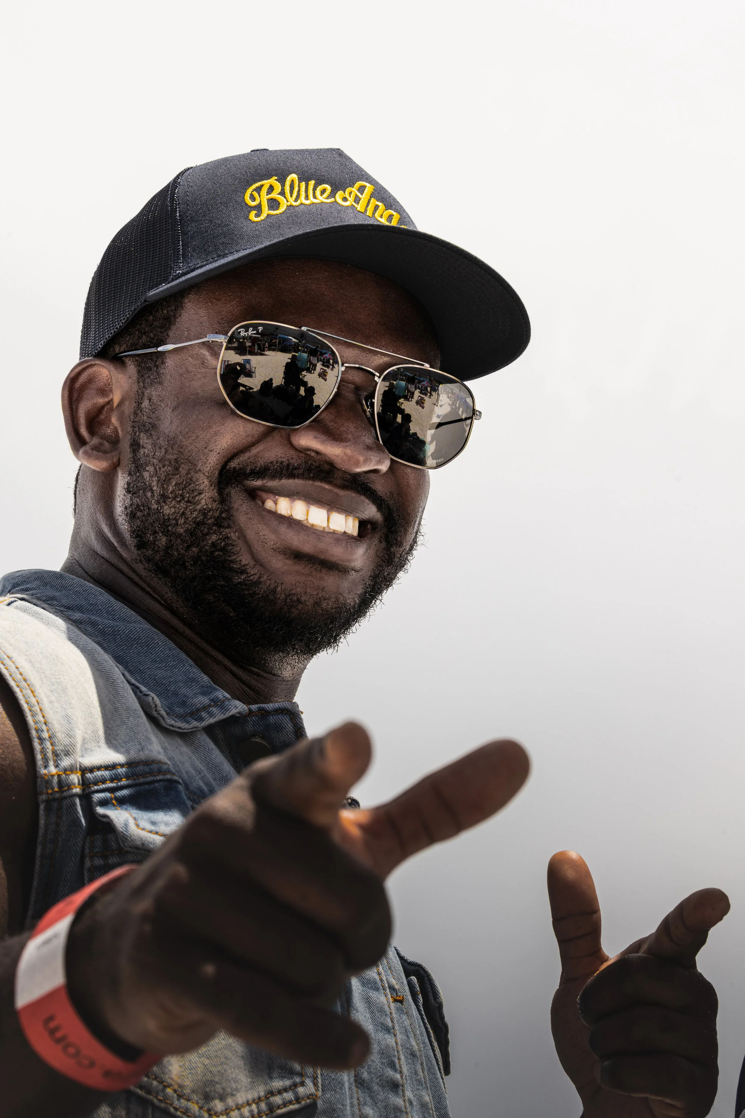 A man smiling, wearing a black cap with yellow lettering, sunglasses, and a denim vest, pointing towards the camera.