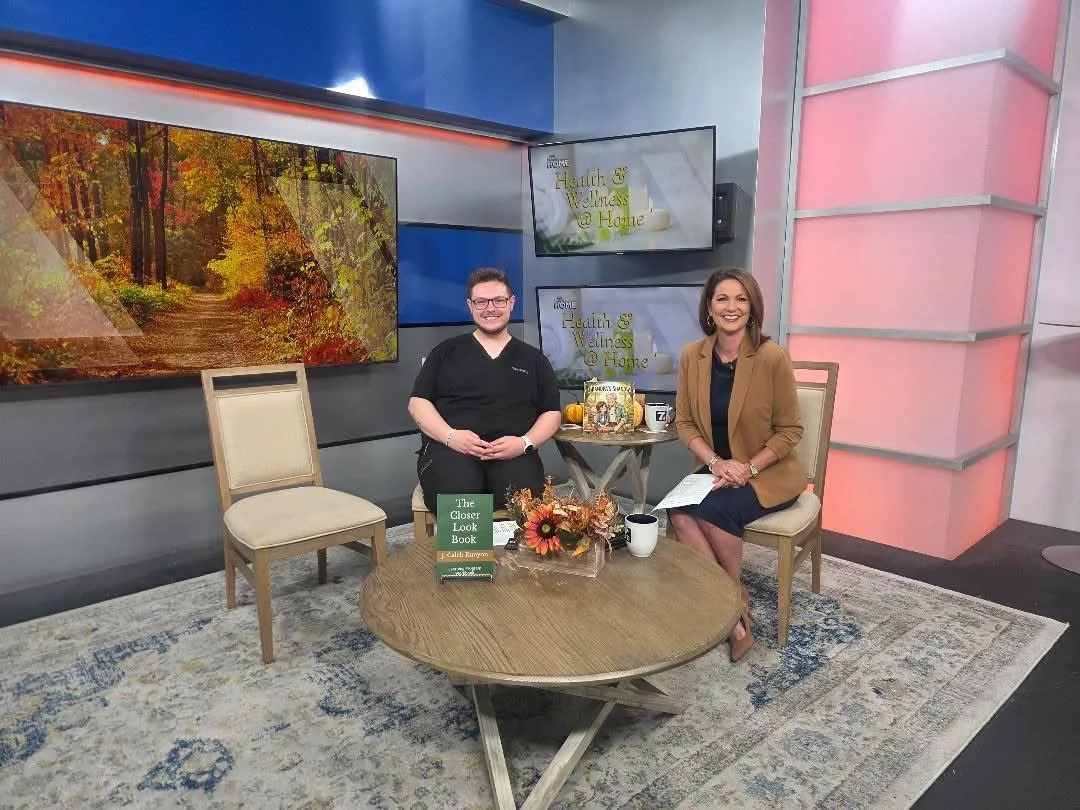 A TV studio set featuring two women sitting at a round table with books, a floral arrangement, and mugs. Behind them are large screens displaying a fall landscape and the show title 'Home Health & Wellness @ Home.'