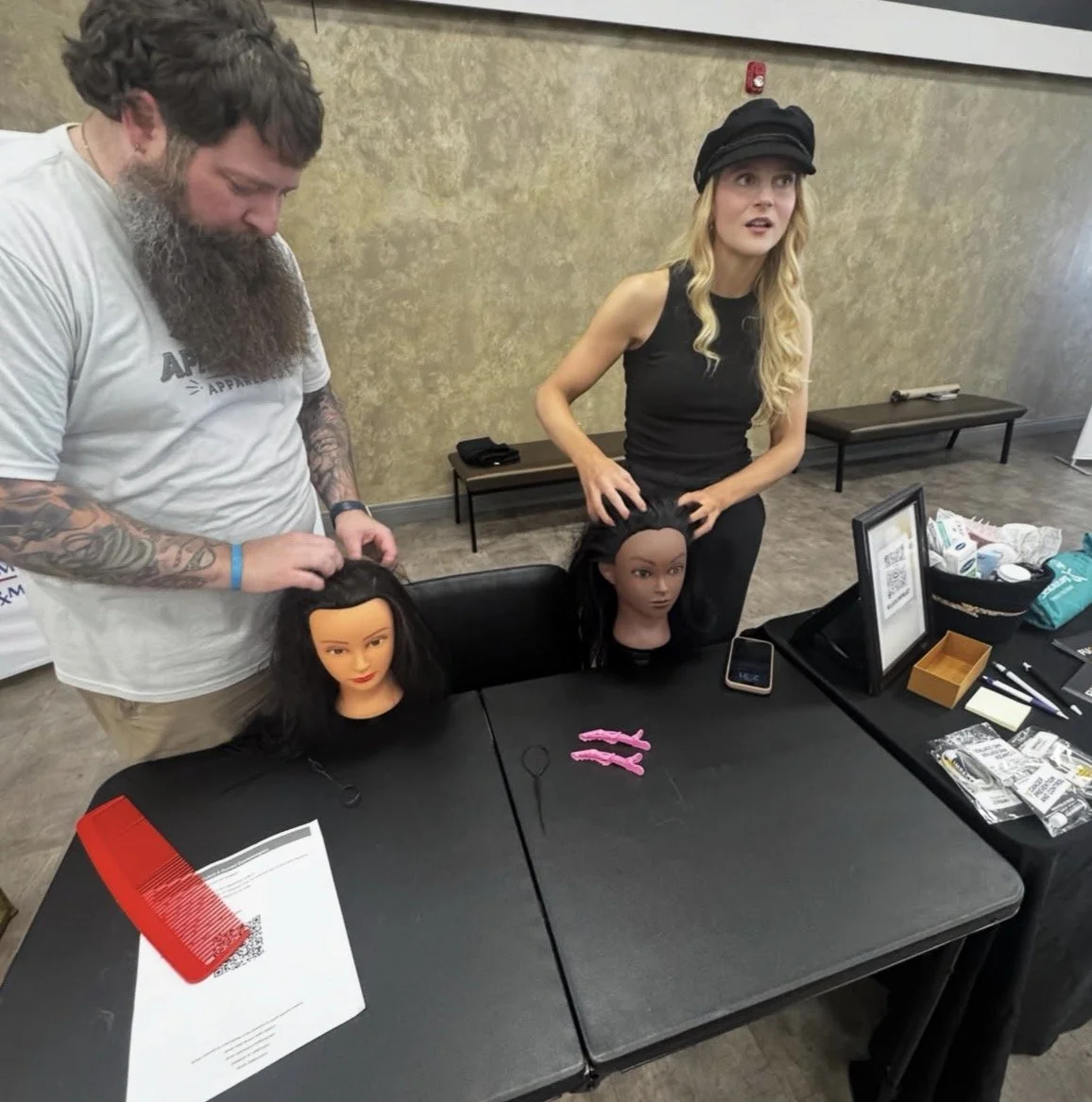 A man stylist with tattoos on his arms styles a mannequin head with black hair, while a woman with long blonde hair and a black hat observes. The scene takes place at a table with hair styling supplies, mannequin heads, and accessories.