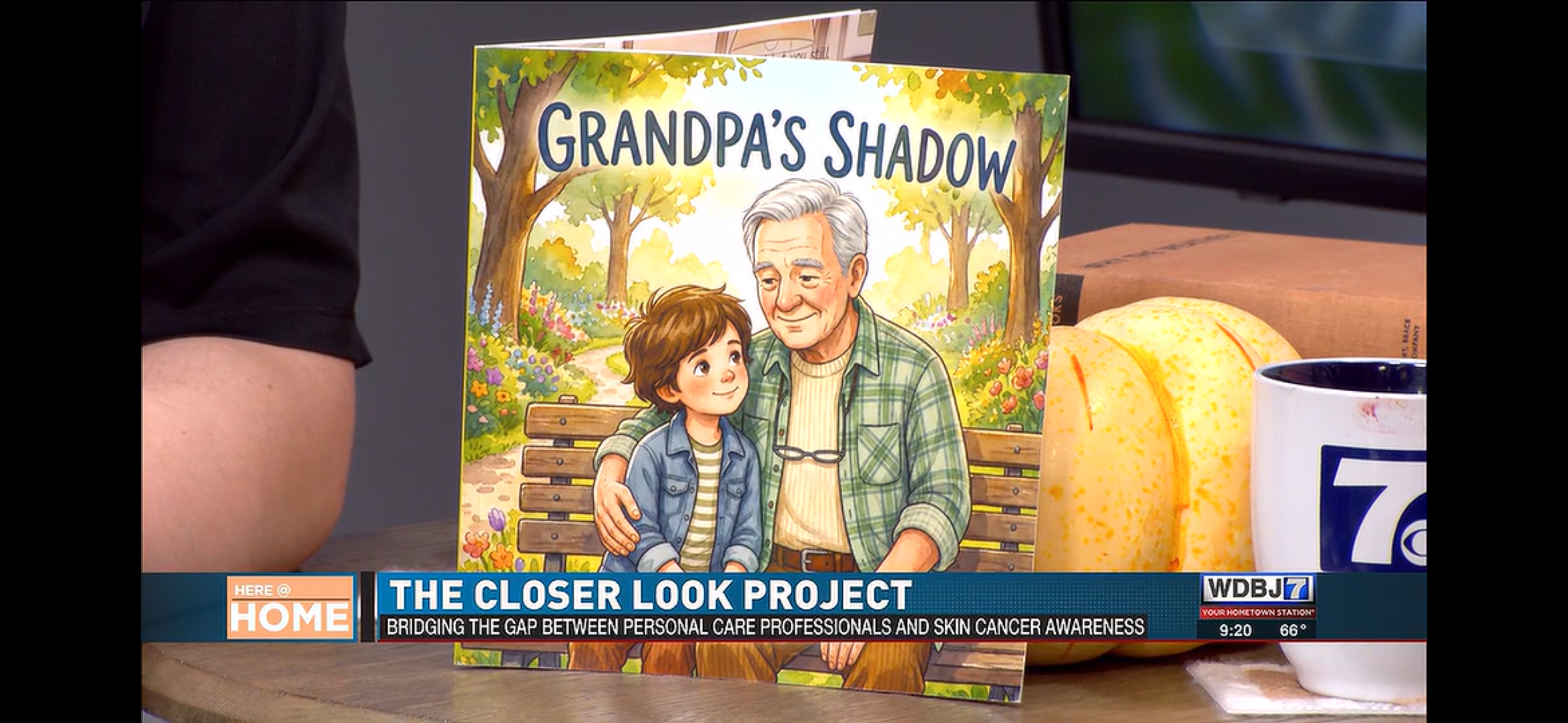 A colorful illustrated children's book titled "Grandpa's Shadow" displayed on a table next to a mug with a yellow pumpkin illustration. The illustration on the book depicts an elderly man and a young boy sitting on a park bench, embracing each other surrounded by trees and flowers.