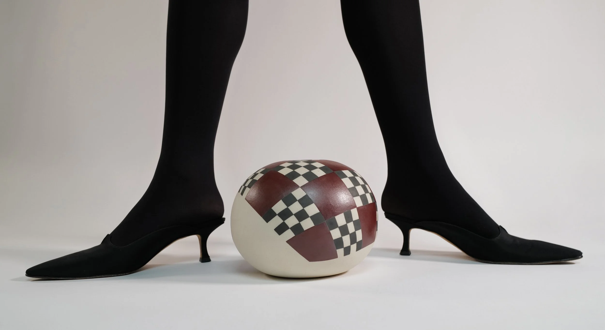 Person standing with feet and legs on either side of a ceramic orb-shaped vessel.  The figure is wearing black tights and pointed-toe heels, and the vessel has a black, maroon and white checkered pattern.
