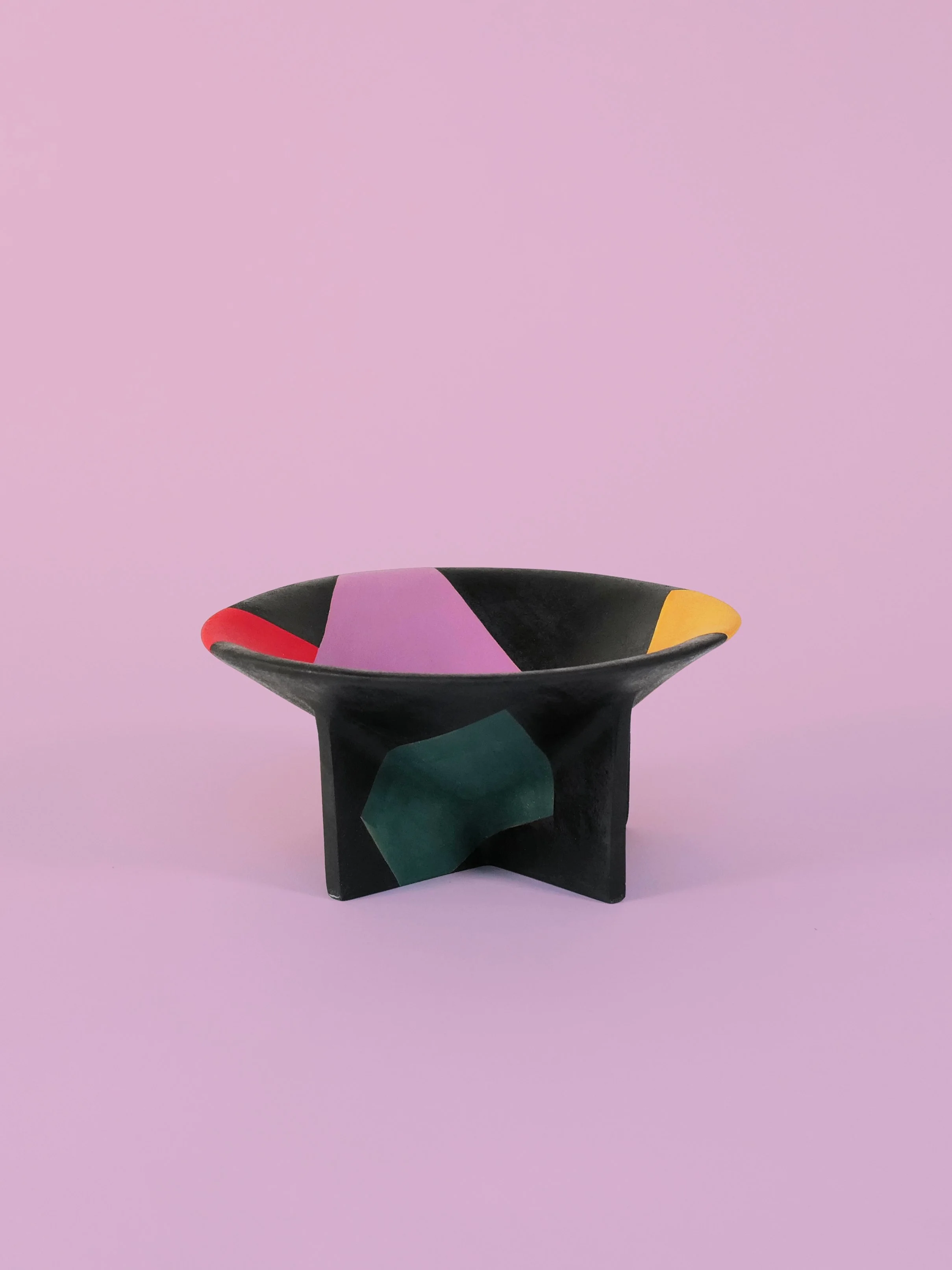 A predominantly black and uniquely shaped ceramic pedestal dish with colorful geometric patterns, placed on a pale purple background.