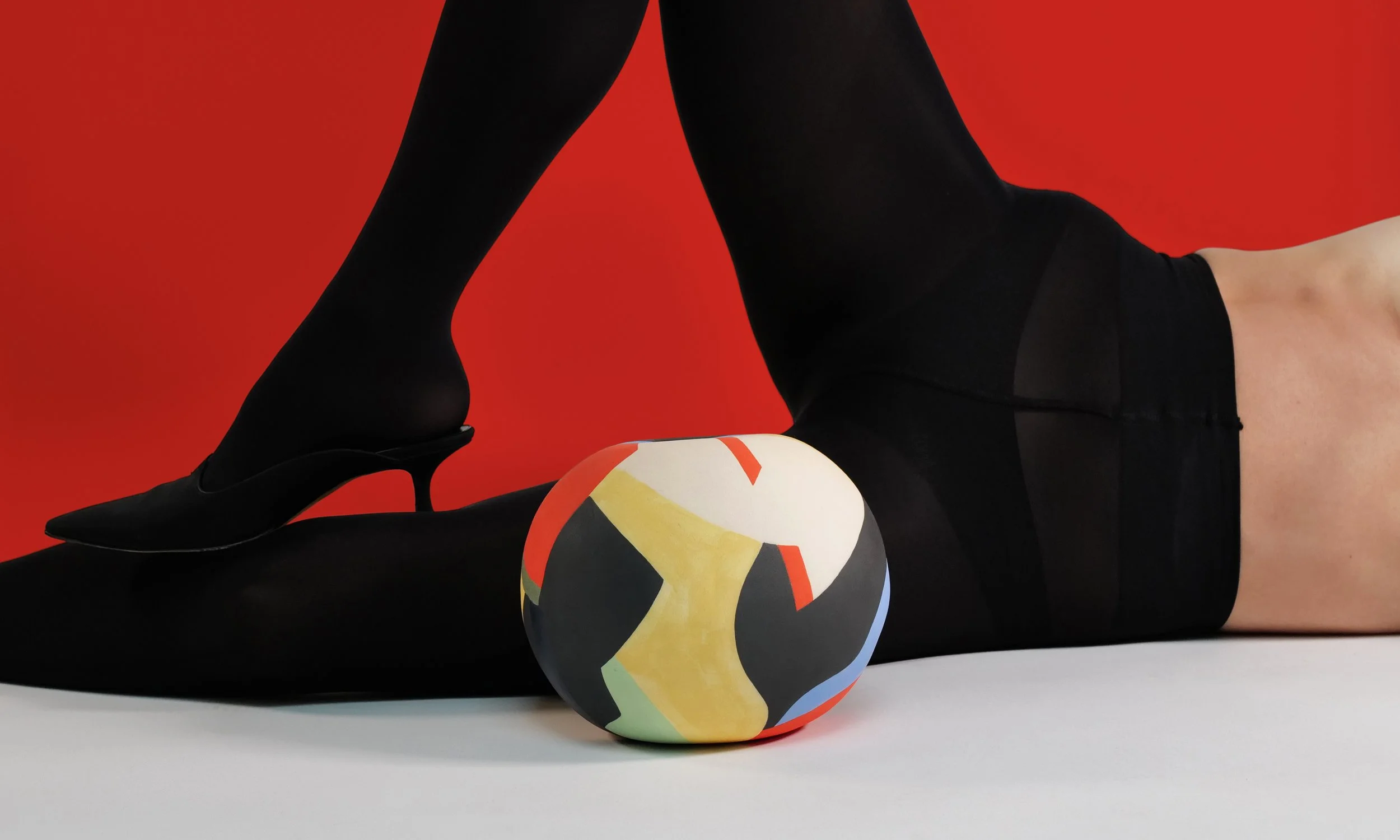 A figure wearing black tights and black shoes lying on the floor against a bold red background with a colorful abstract-patterned ceramic vessel in front of them.