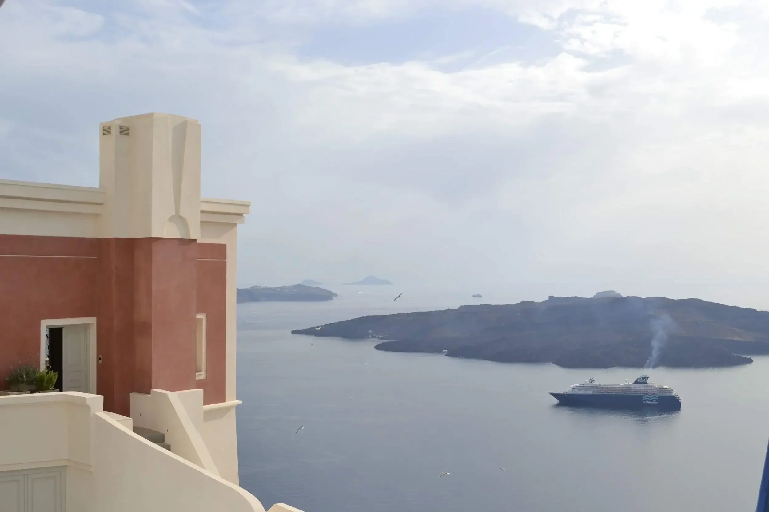 View of a cruise ship sailing near a small island seen from a building with white and pink walls, overlooking the ocean on a cloudy day.