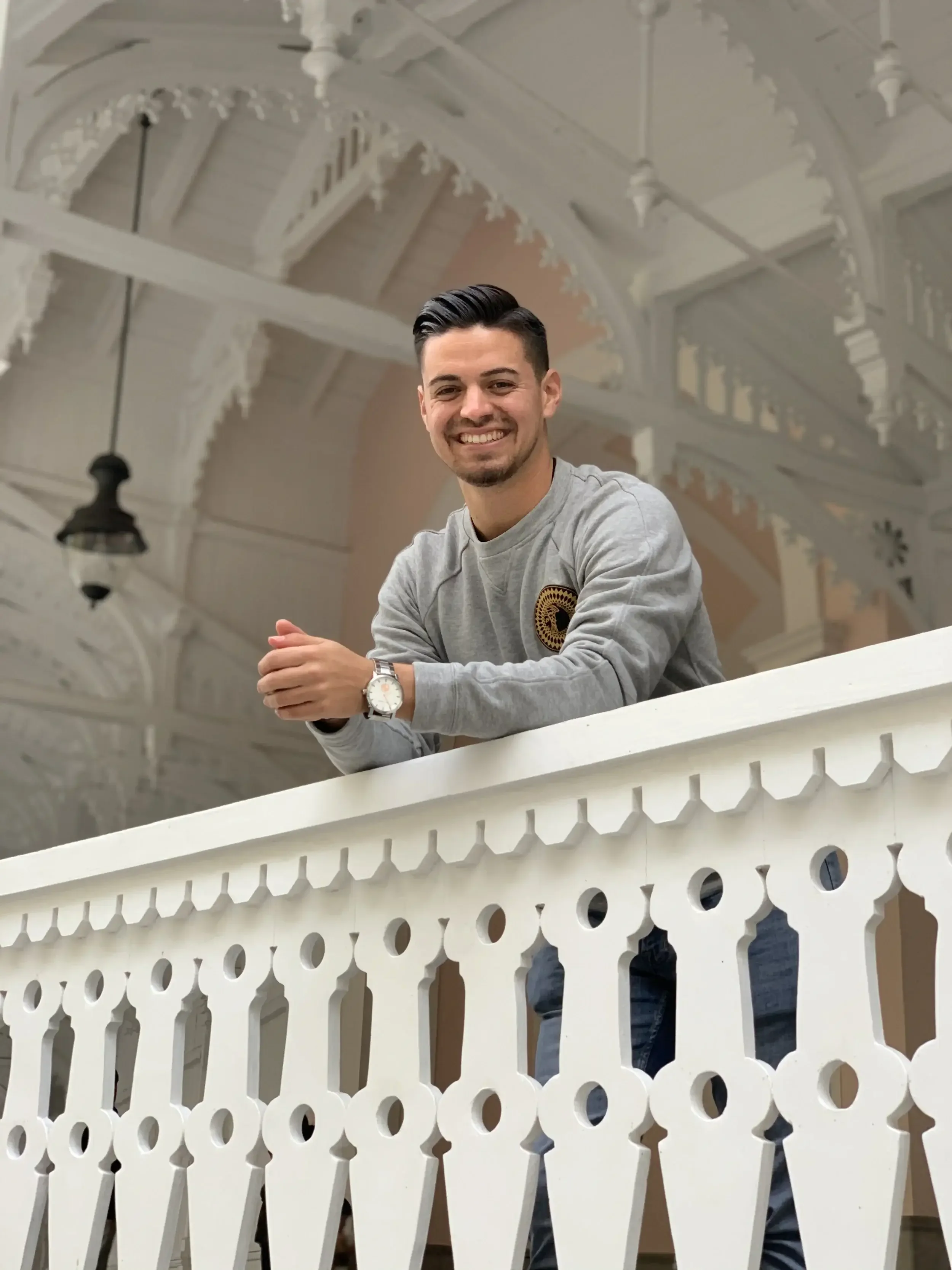 A young man with dark hair styled back, wearing a gray sweatshirt with a circular logo on the chest, smiling and leaning on a white decorative railing inside a building with ornate white wooden architecture and a hanging light fixture in the background.