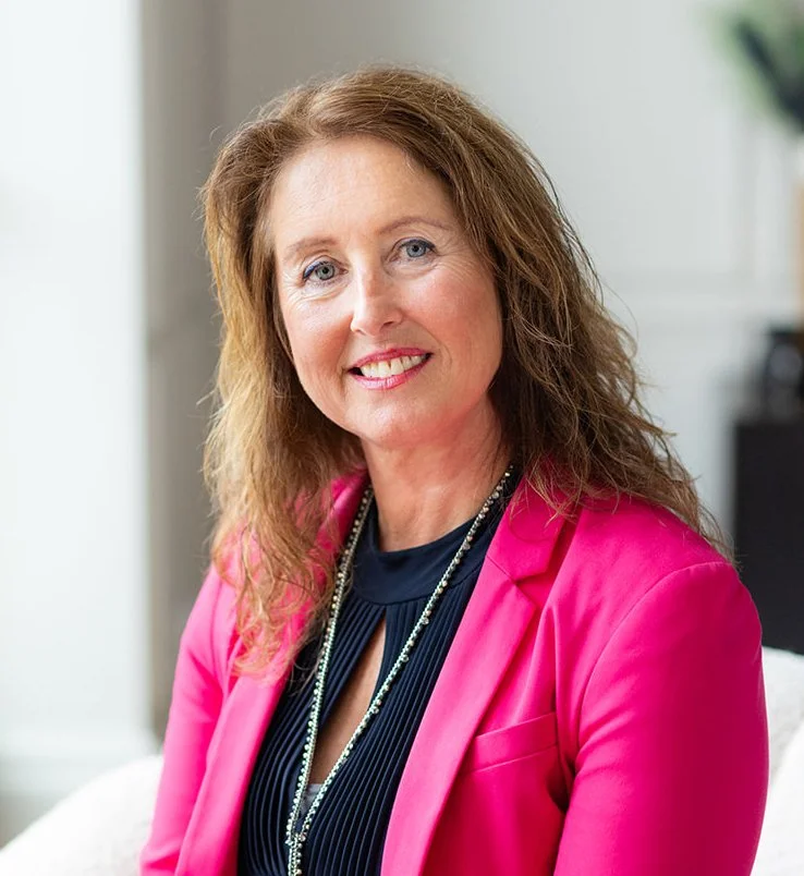 Susan Foulger, smiling, with shoulder-length red hair, blue eyes, wearing a pink blazer over a dark top, with a beaded necklace, seated in a bright room.