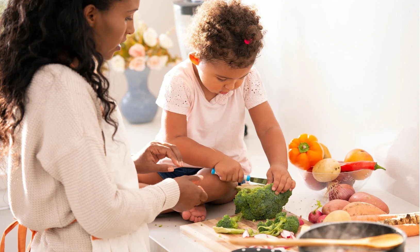 A young girl learning how to cook vegetables with an adult woman, peeling broccoli in a bright kitchen with a bowl of colourful vegetables in the background.