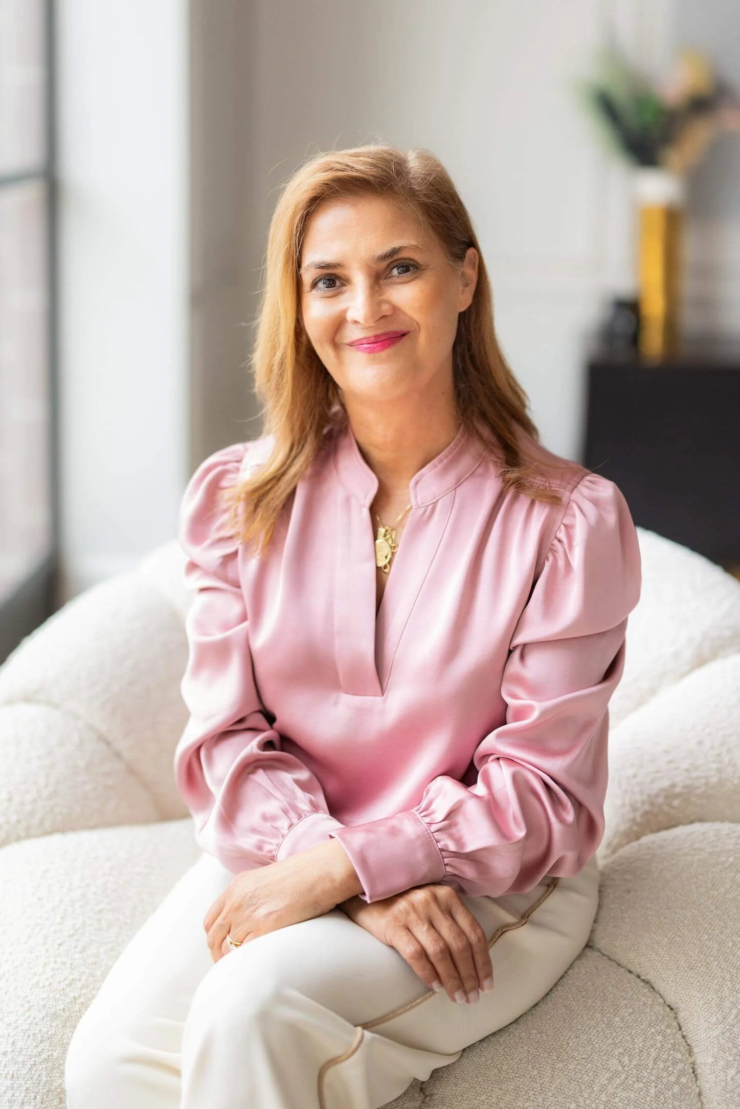 Stella wearing a pink satin blouse sitting on a beige couch inside a well-lit room with light-colored walls, blurred bookshelf or decor in the background.