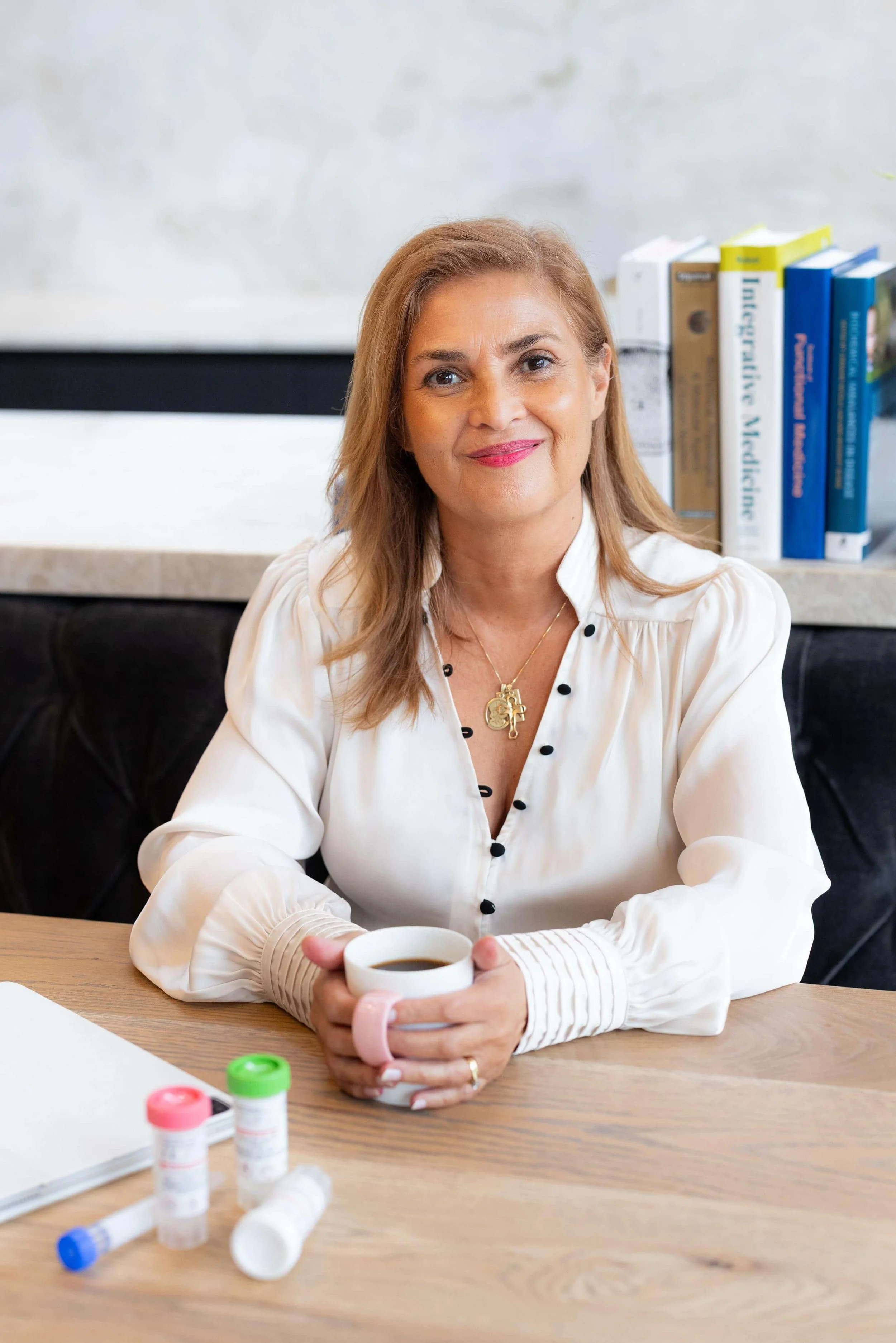 Stella in a white blouse, sitting at a wooden table, holding a mug of coffee, with pills and medication bottles on the table, and a bookshelf with books behind her.