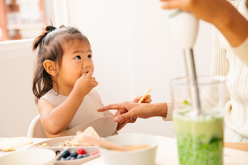 A young girl with dark hair in a ponytail eating breakfast while a woman points at her, while making a green juice drink.