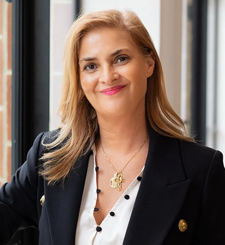 Stella Chadwick, founder of Brainstorm Health, wearing a black blazer and a white blouse, smiling, standing near a large window.