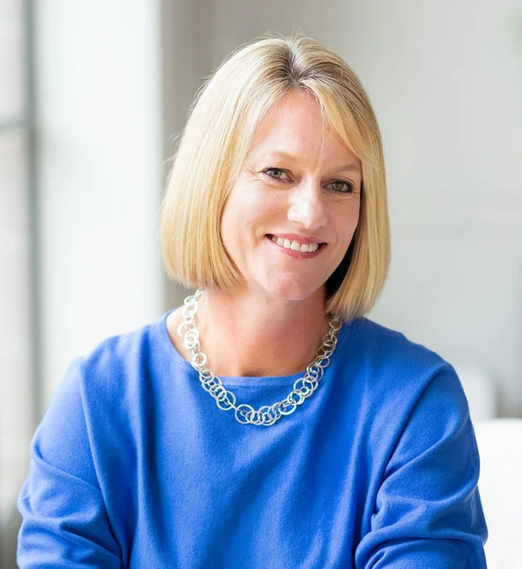 Carolyn Frost, Nutritional Therapist and Homeopath, with shoulder-length blonde hair, wearing a blue top and a silver chain necklace, smiling at the camera in a well-lit indoor setting.