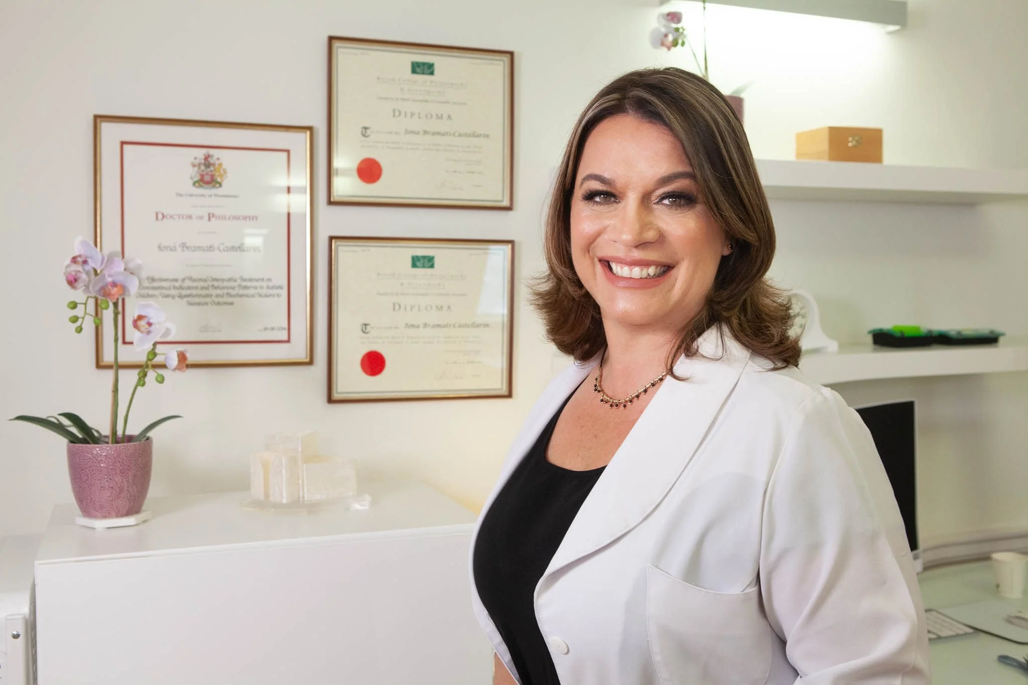 Dr Ioná Bramati, a woman with shoulder-length brown hair smiling, wearing a white blazer over a black top, standing in a clinic with diplomas and certificates on the wall behind her, and a potted orchid on a white counter.