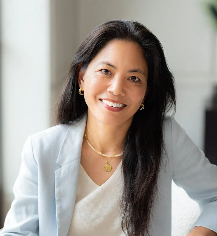 Mémé Watanabe, a woman with long black hair smiling, wearing a white blazer and gold jewellery, sitting indoors.